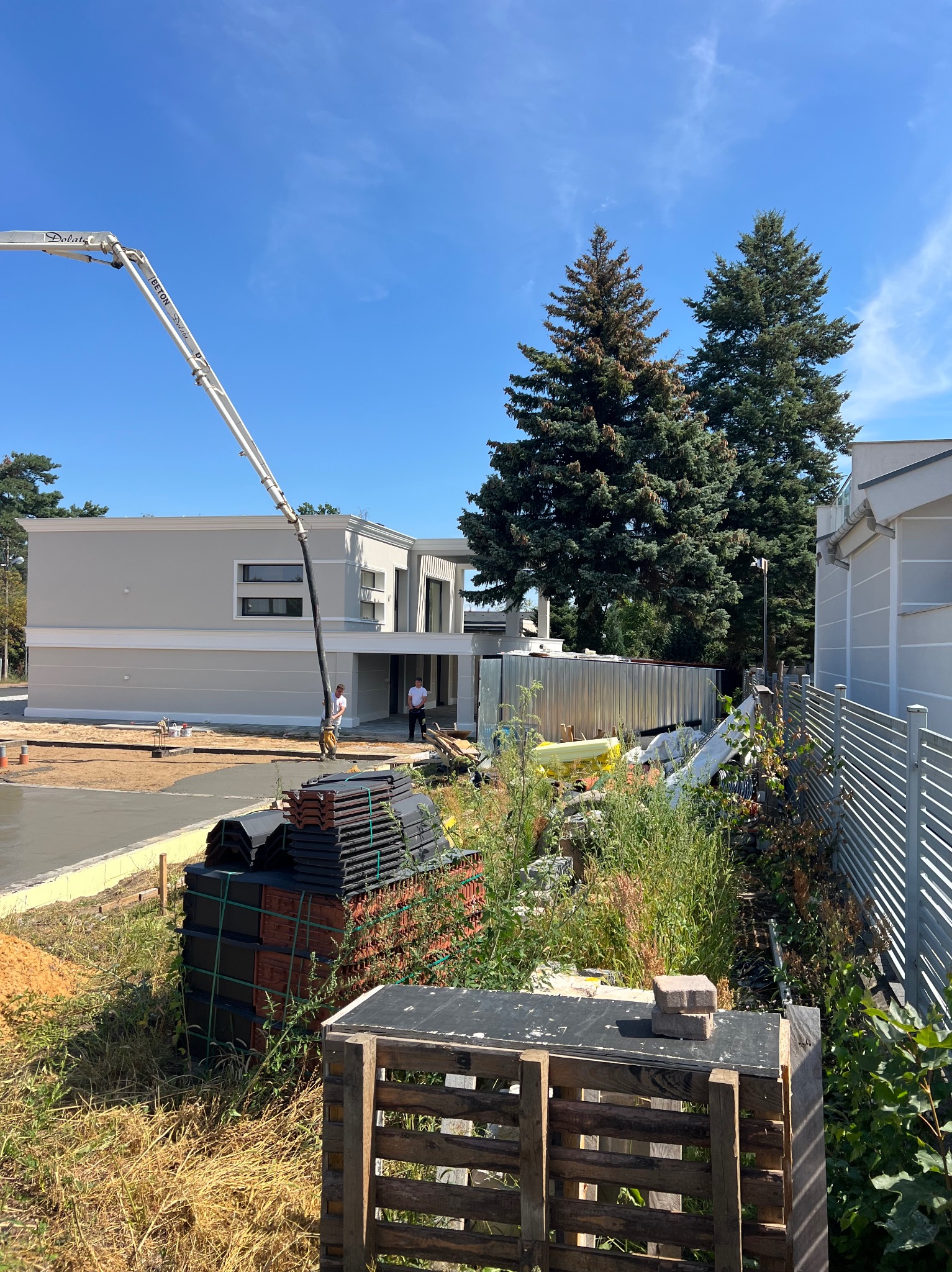 Wylewka betonowa przed nowoczesnym domem z użyciem pompy do betonu. Na pierwszym planie paleta z dachówkami. Słoneczny dzień, błękitne niebo.