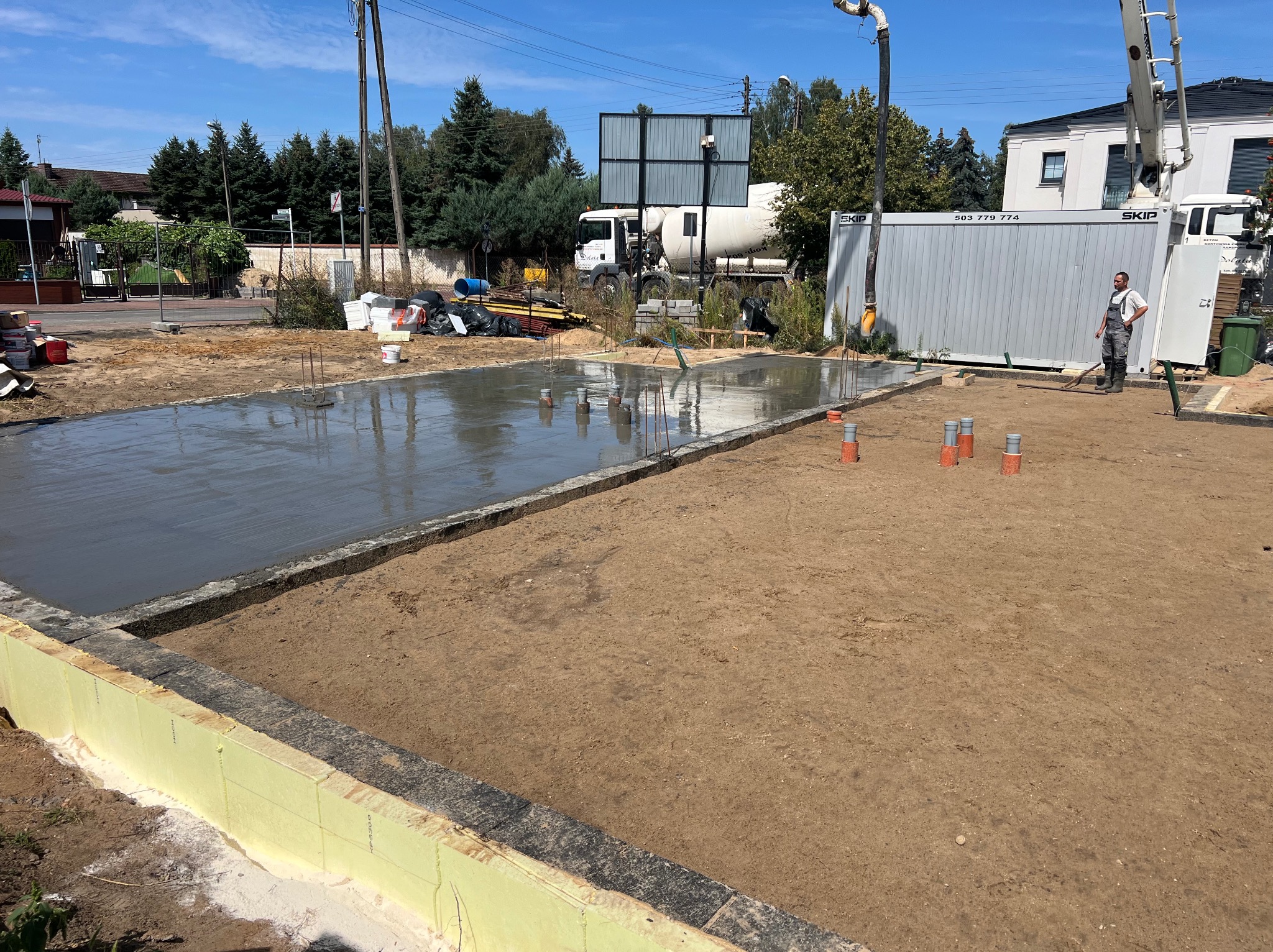 Wylany fundament pod dom jednorodzinny z widoczną izolacją, rurami kanalizacyjnymi i pompą do betonu. W tle betoniarka i kontener budowlany.