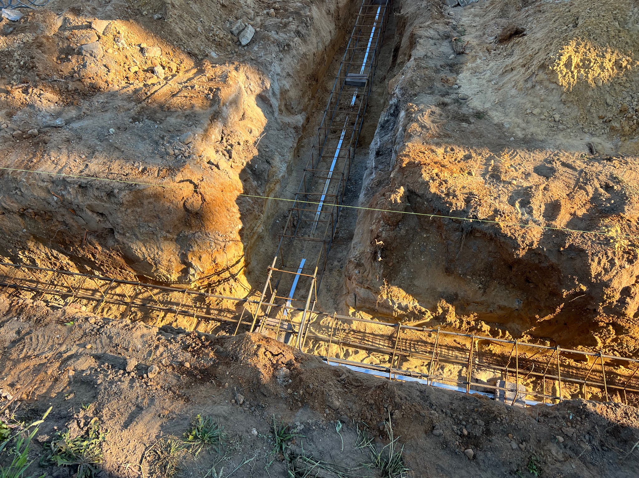 Wykop z ułożonym zbrojeniem i rurami kanalizacyjnymi, przygotowany do zalania fundamentów. Widok z góry, oświetlony popołudniowym słońcem.