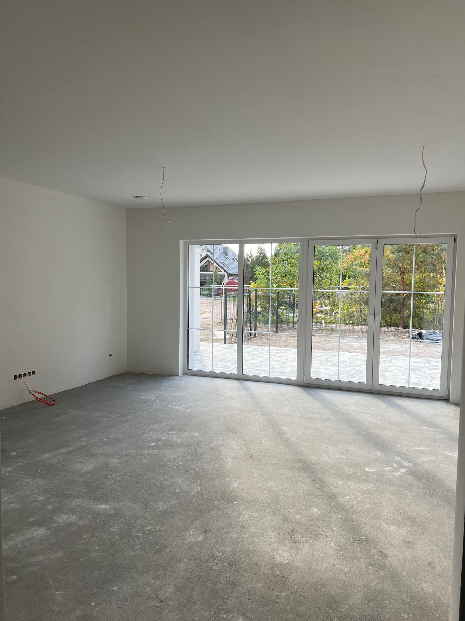 Surowe wnętrze z dużym oknem tarasowym i widocznymi instalacjami elektrycznymi. Szara, betonowa podłoga i białe ściany w nowym domu.