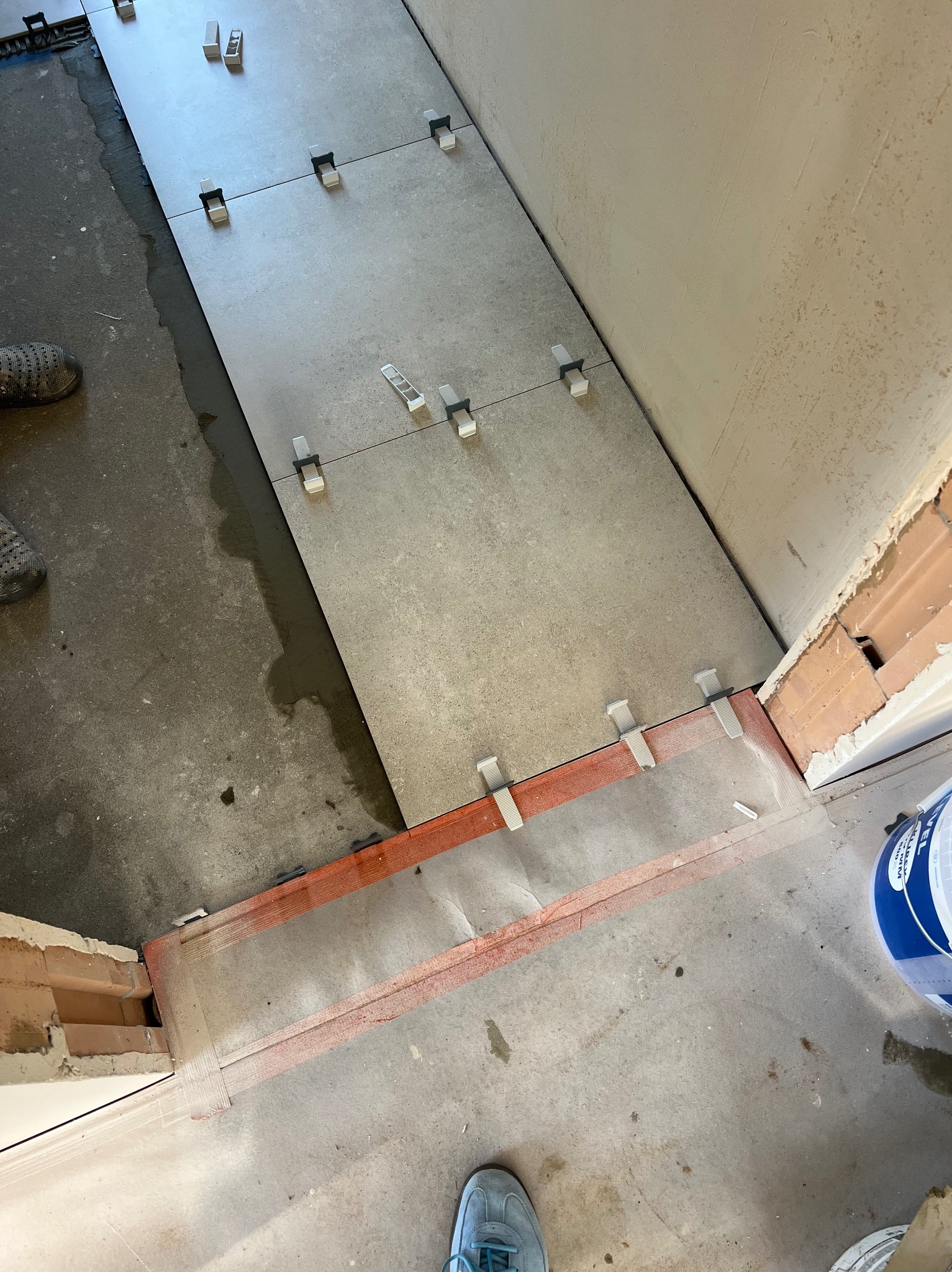 Układanie szarych płytek podłogowych z użyciem klipsów poziomujących. Widoczne fragmenty butów i wiadro z zaprawą. Prace wykończeniowe w trakcie realizacji.