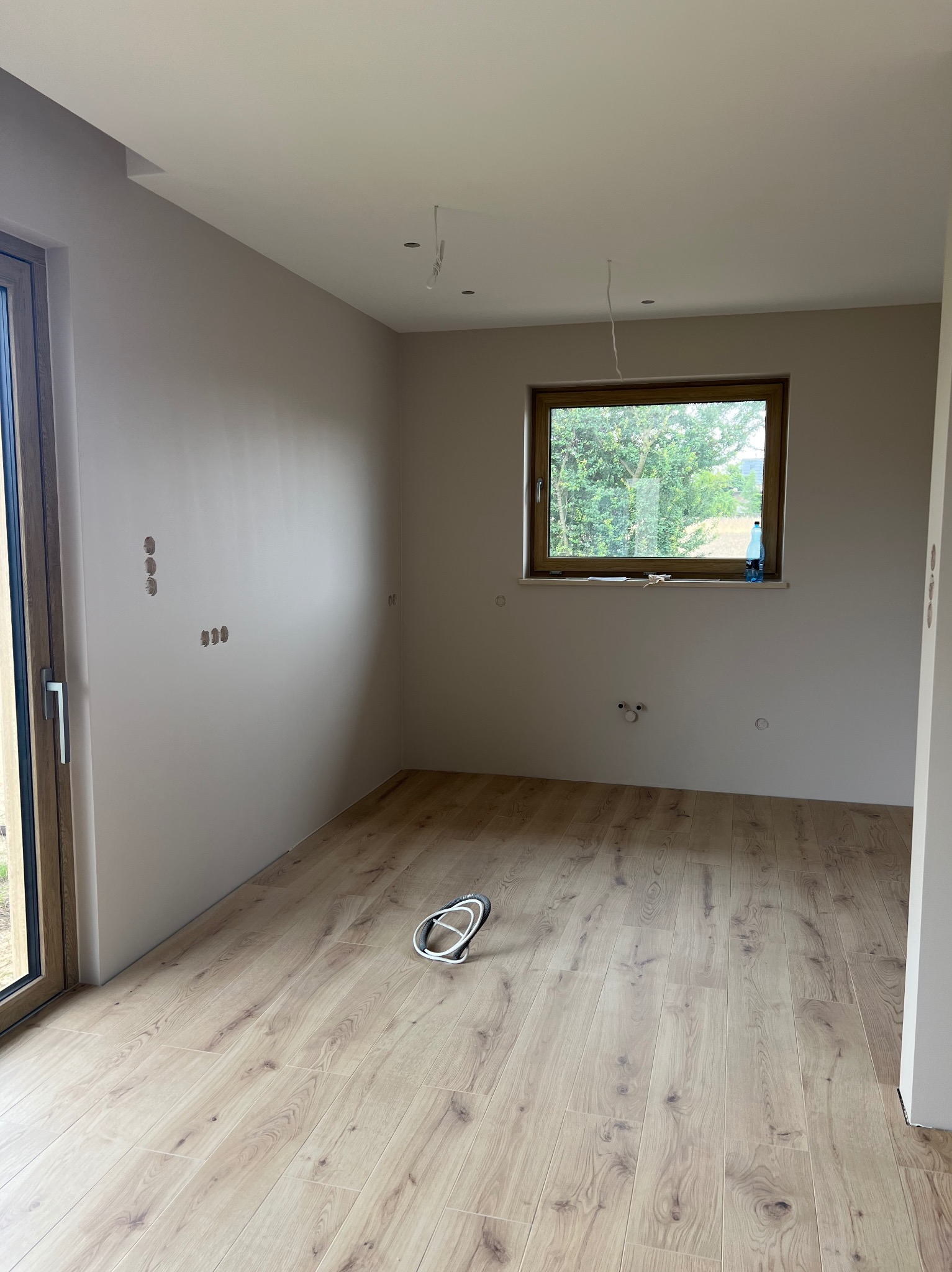 Jasne panele podłogowe w pokoju z beżowymi ścianami, widoczne puszki elektryczne, okno z widokiem na zieleń i kabel od oświetlenia sufitowego. Wykończenie wnętrz w nowym domu.