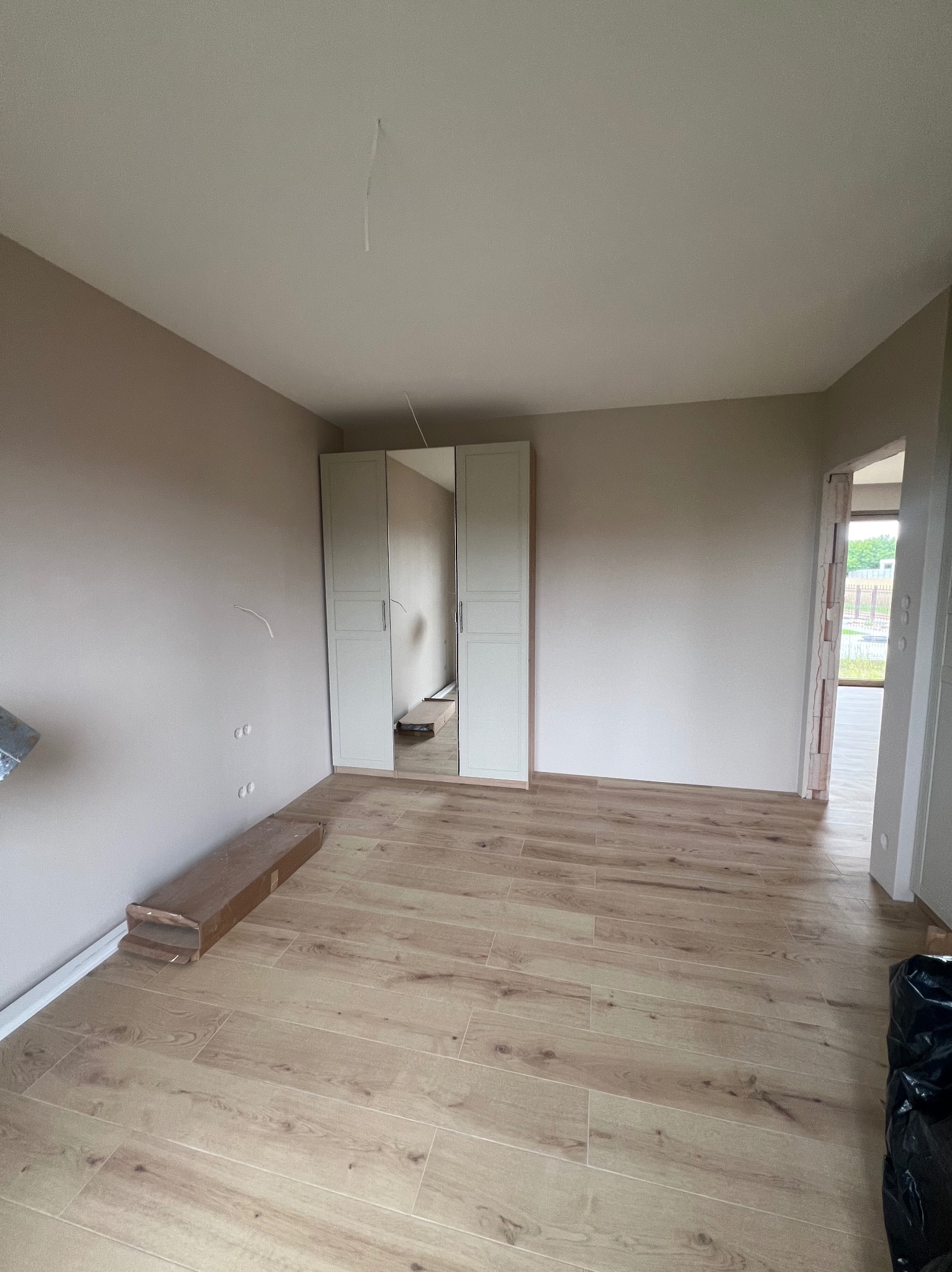 Jasne panele podłogowe w pokoju z beżowymi ścianami i białą szafą z lustrem. Widoczne niedokończone detale, np. wystające kable i listwy przypodłogowe.
