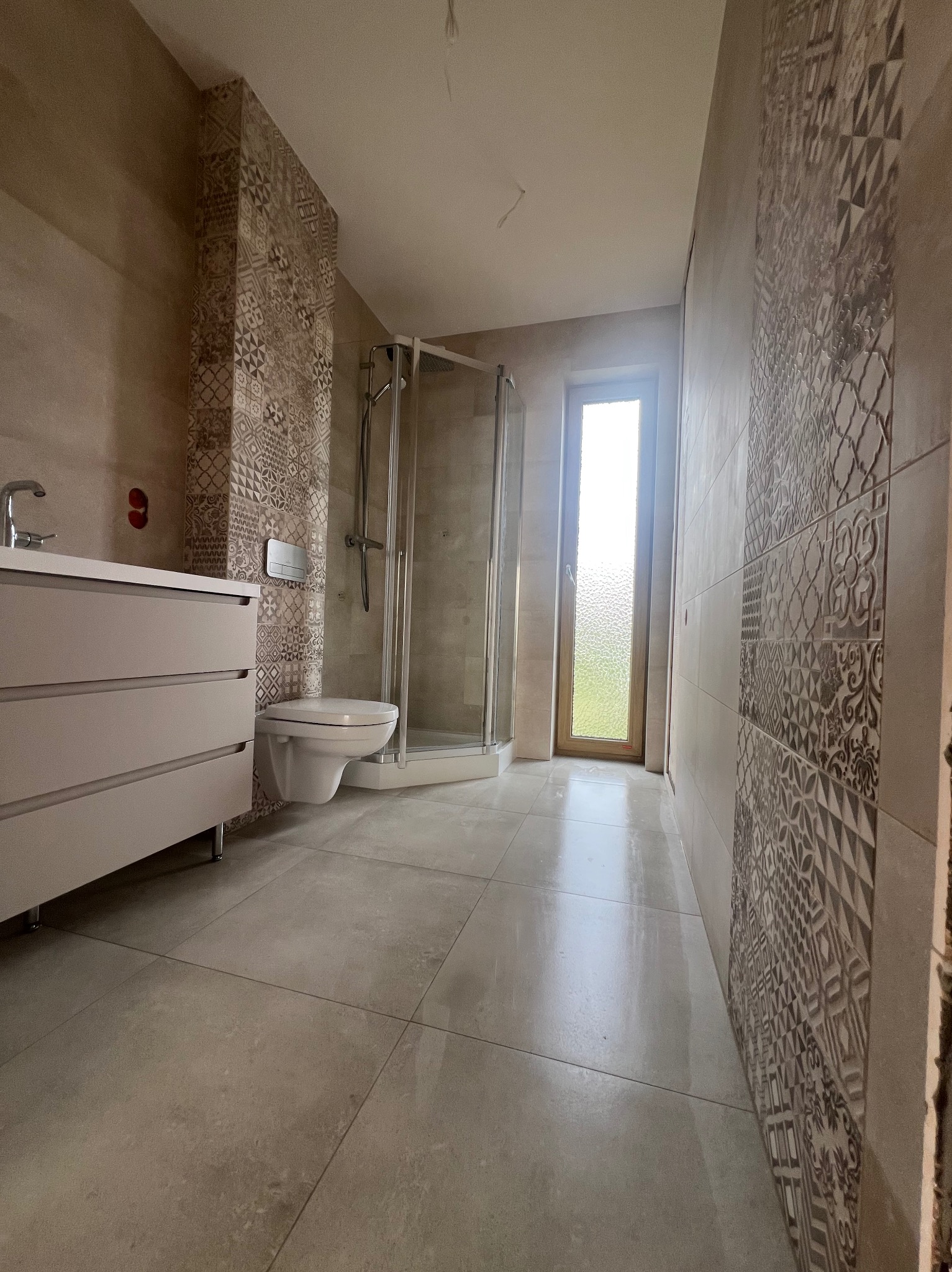 Jasna łazienka z kabiną prysznicową, umywalką z szafką i toaletą podwieszaną. Ściany wyłożone płytkami z wzorem, podłoga z dużych, jasnych płytek. Okno w tle.