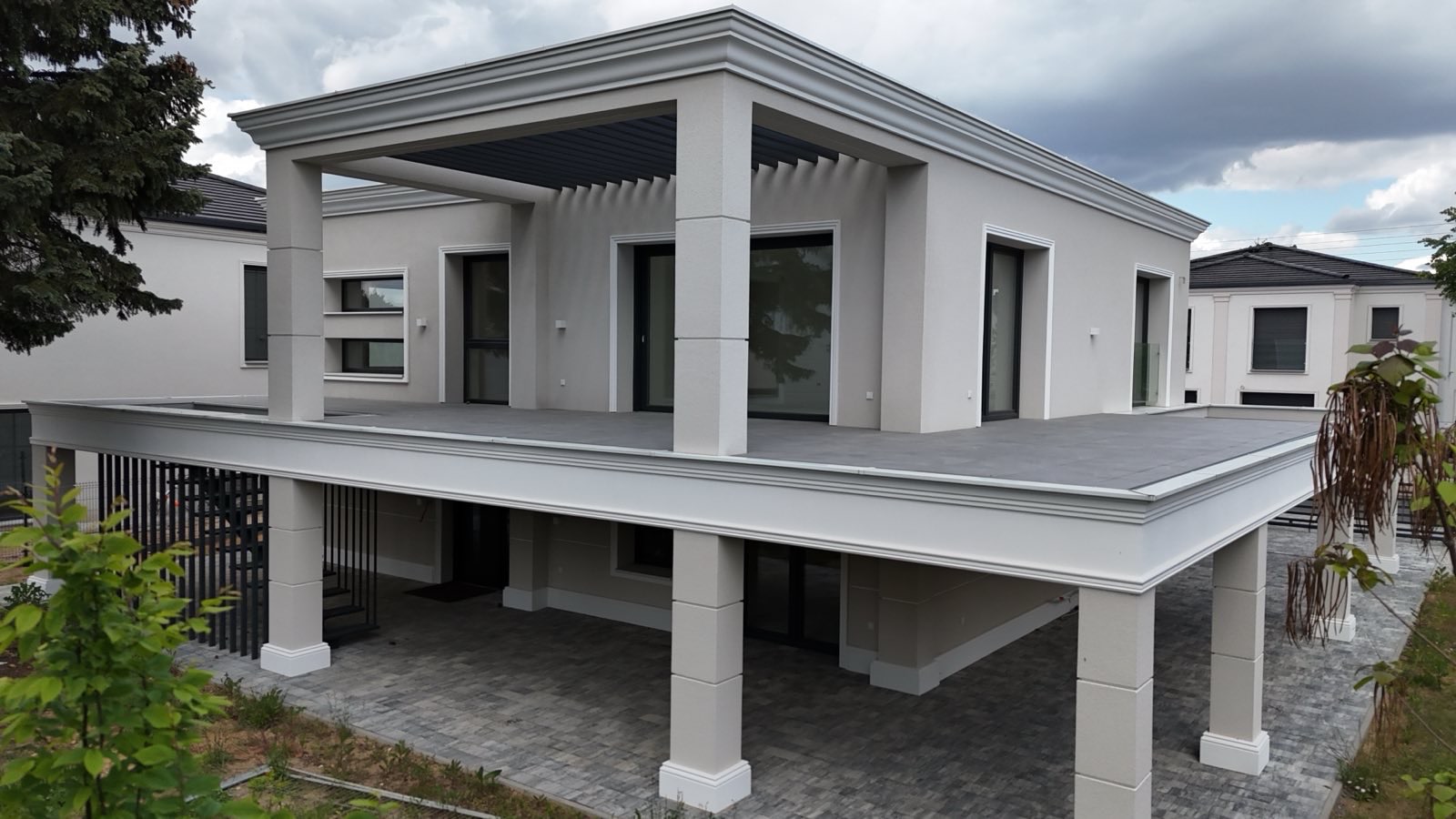 Nowoczesny dom z płaskim dachem i kolumnami, wykończony w jasnych barwach. Widoczny taras na piętrze i starannie ułożona kostka brukowa na podjeździe.