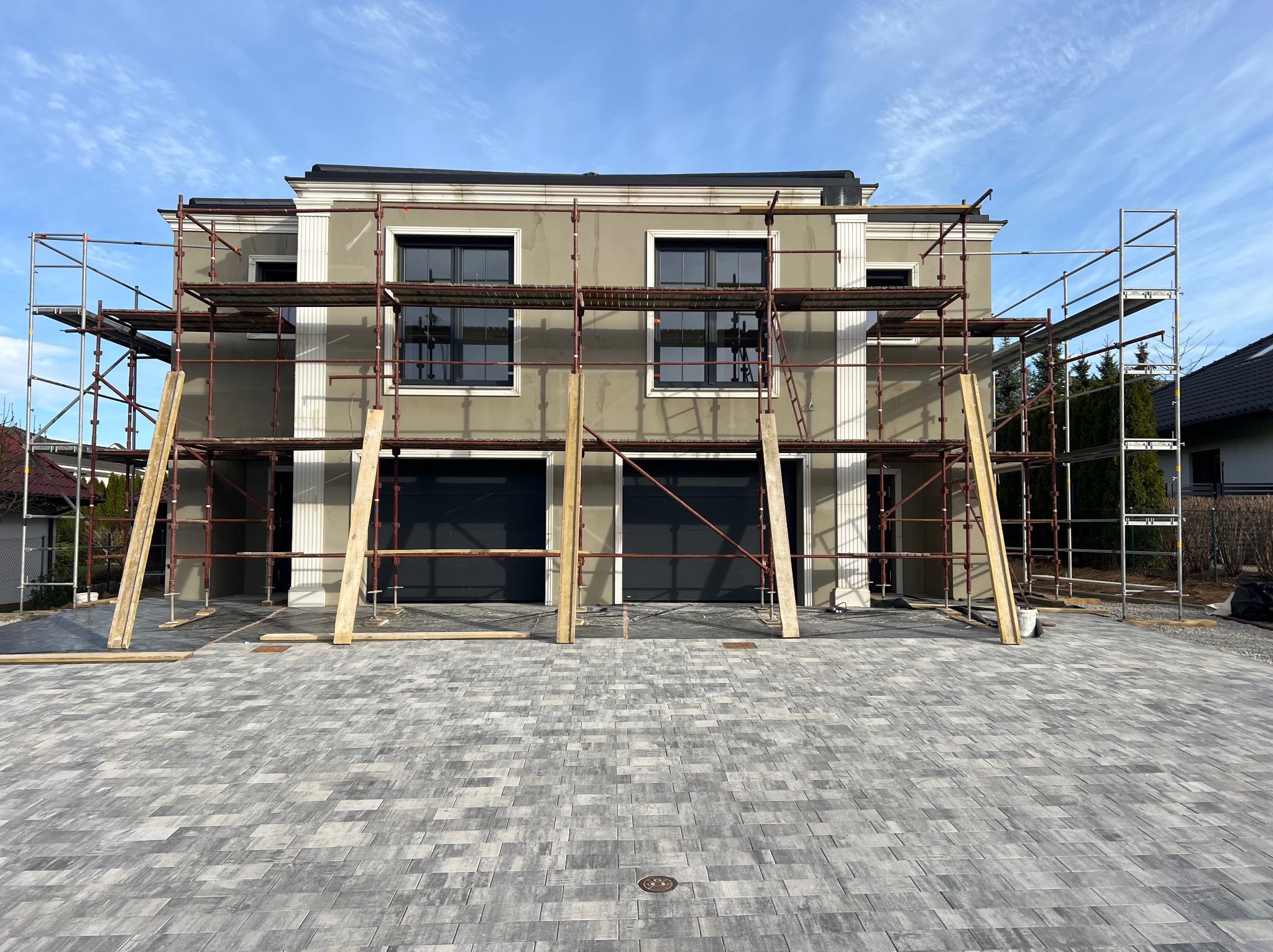 Dwukondygnacyjny dom w trakcie elewacji, otoczony rusztowaniami. Widoczne szare ściany, czarne okna i brama garażowa. Brukowany podjazd i błękitne niebo w tle.