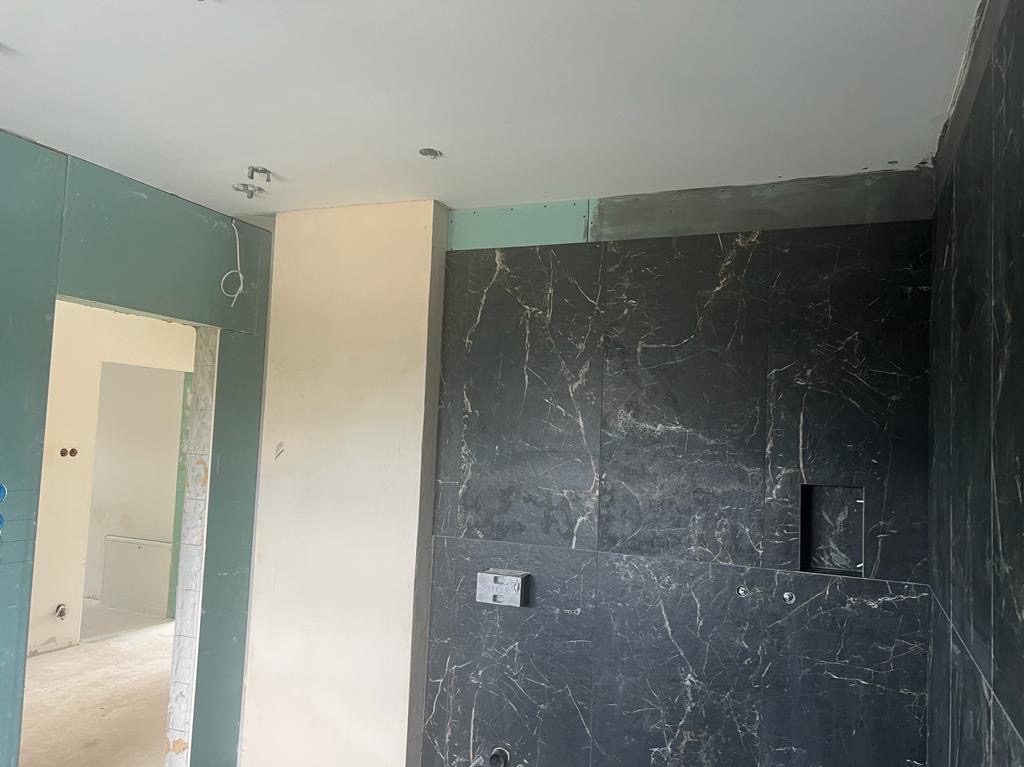 Łazienka w trakcie wykończenia: czarne płytki imitujące marmur na ścianie, surowa płyta gipsowa na ścianach, widoczne instalacje. Wnętrze w stanie deweloperskim.