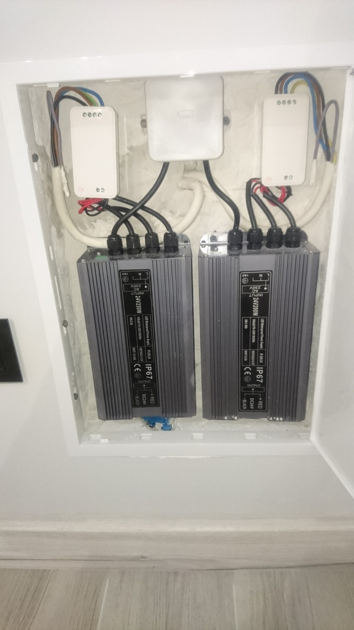 Dwa zasilacze LED w białej wnęce, widoczne przewody i moduły sterujące. Instalacja elektryczna w trakcie montażu, detale połączeń i specyfikacja techniczna zasilaczy.