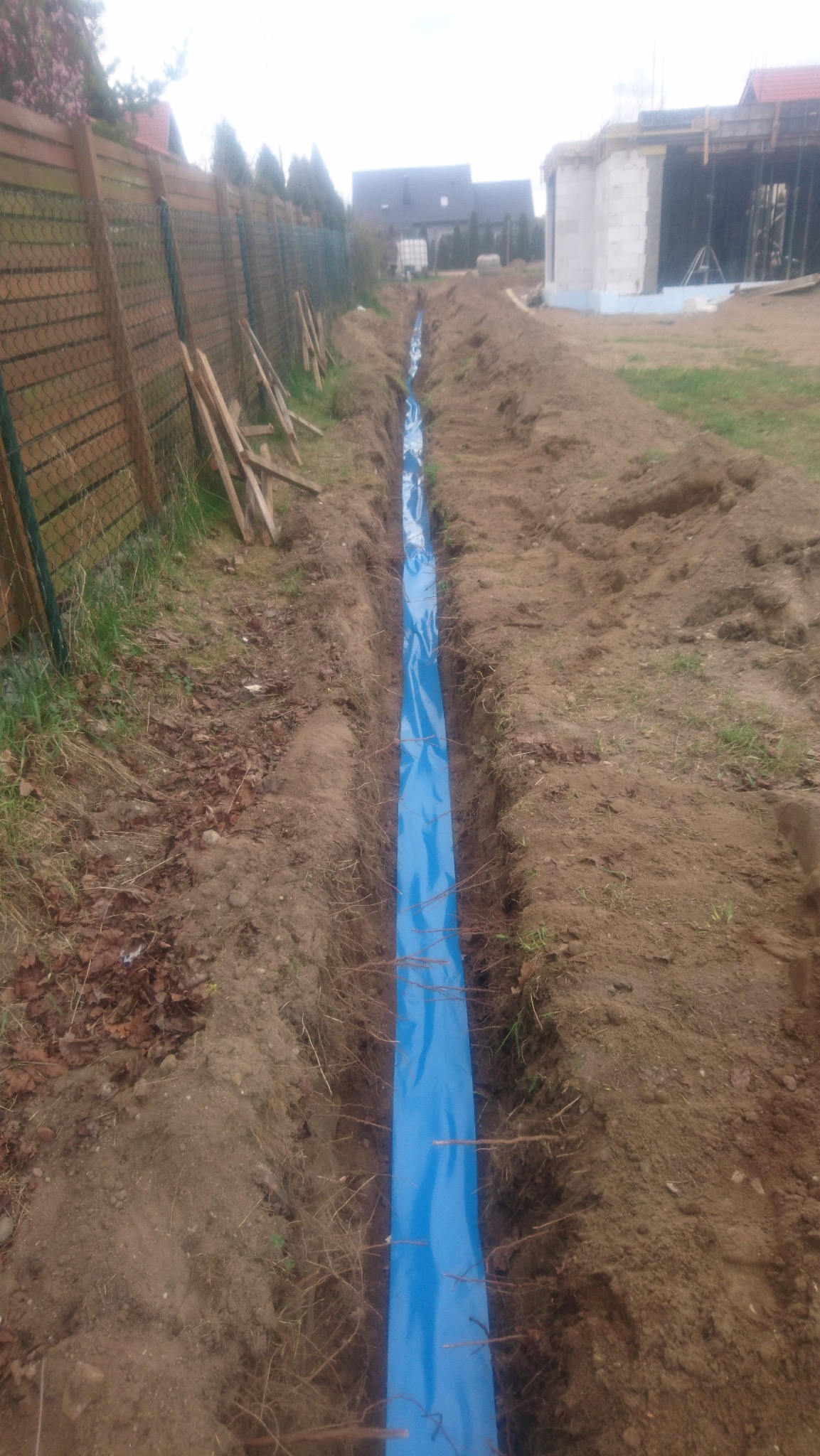 Wykop z niebieską folią, prawdopodobnie pod instalację odgromową. Widoczne wystające pręty i ziemia. W tle budynek w trakcie budowy i ogrodzenie.
