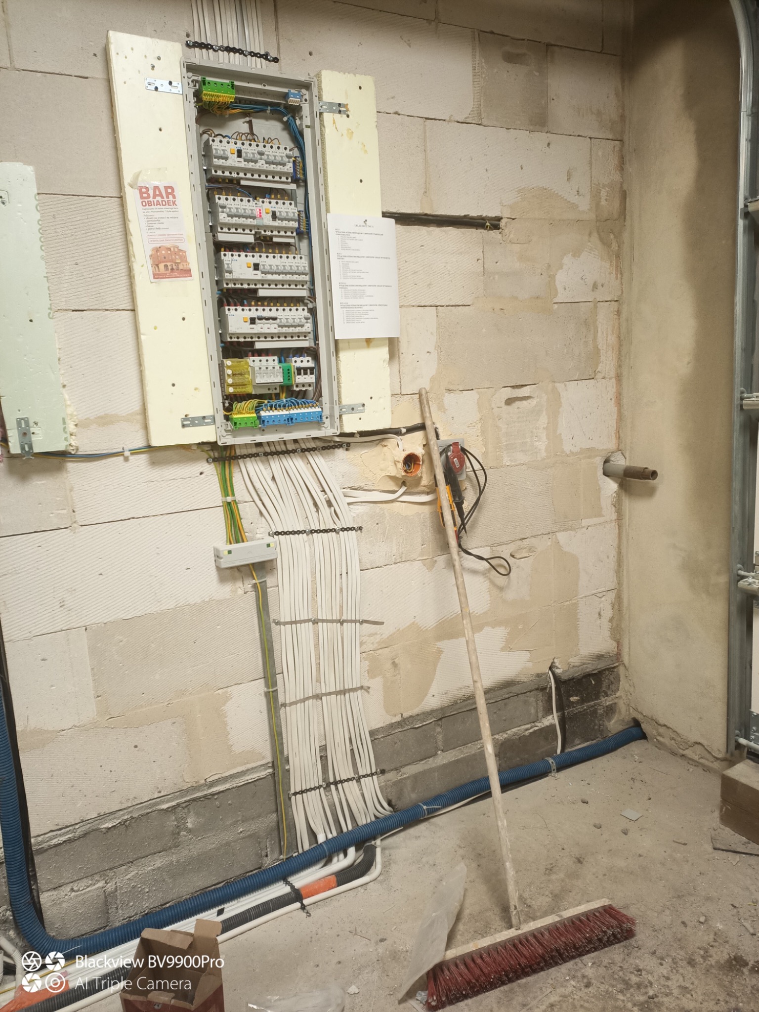 Rozdzielnia elektryczna z licznymi przewodami na ścianie z betonu komórkowego. Widoczne zabezpieczenia, listwa zaciskowa i kable. W tle ściana w surowym stanie.