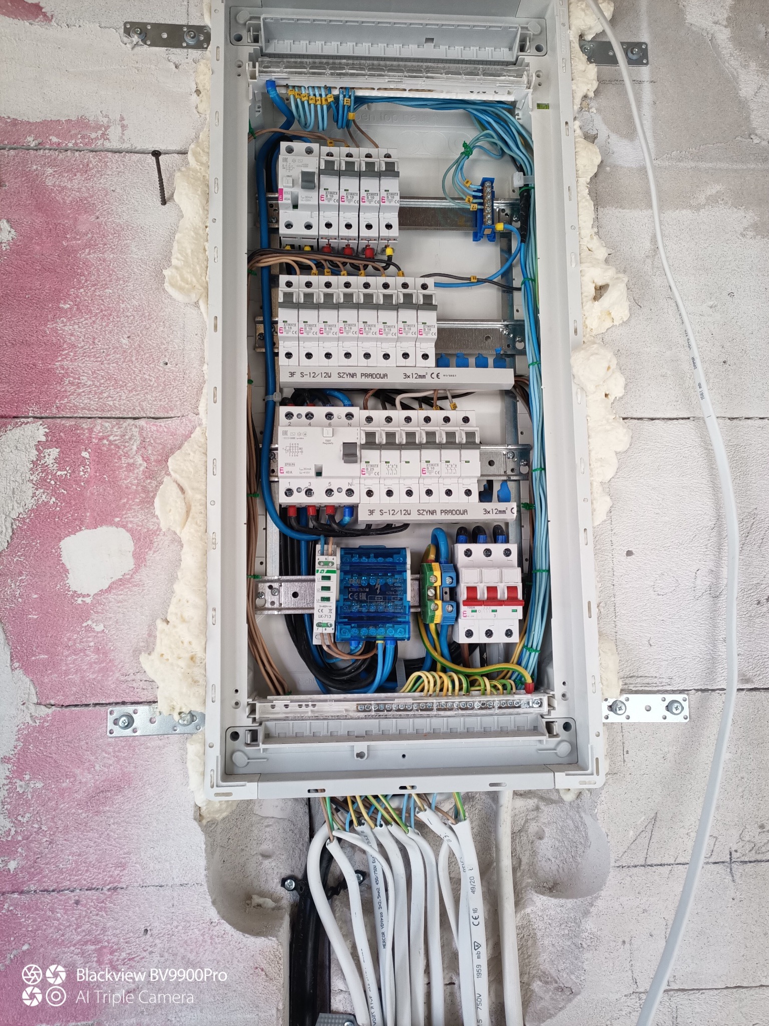 Rozdzielnica elektryczna wnękowa, zamontowana w surowej ścianie z widocznymi przewodami. Widoczne zabezpieczenia, bezpieczniki i okablowanie.