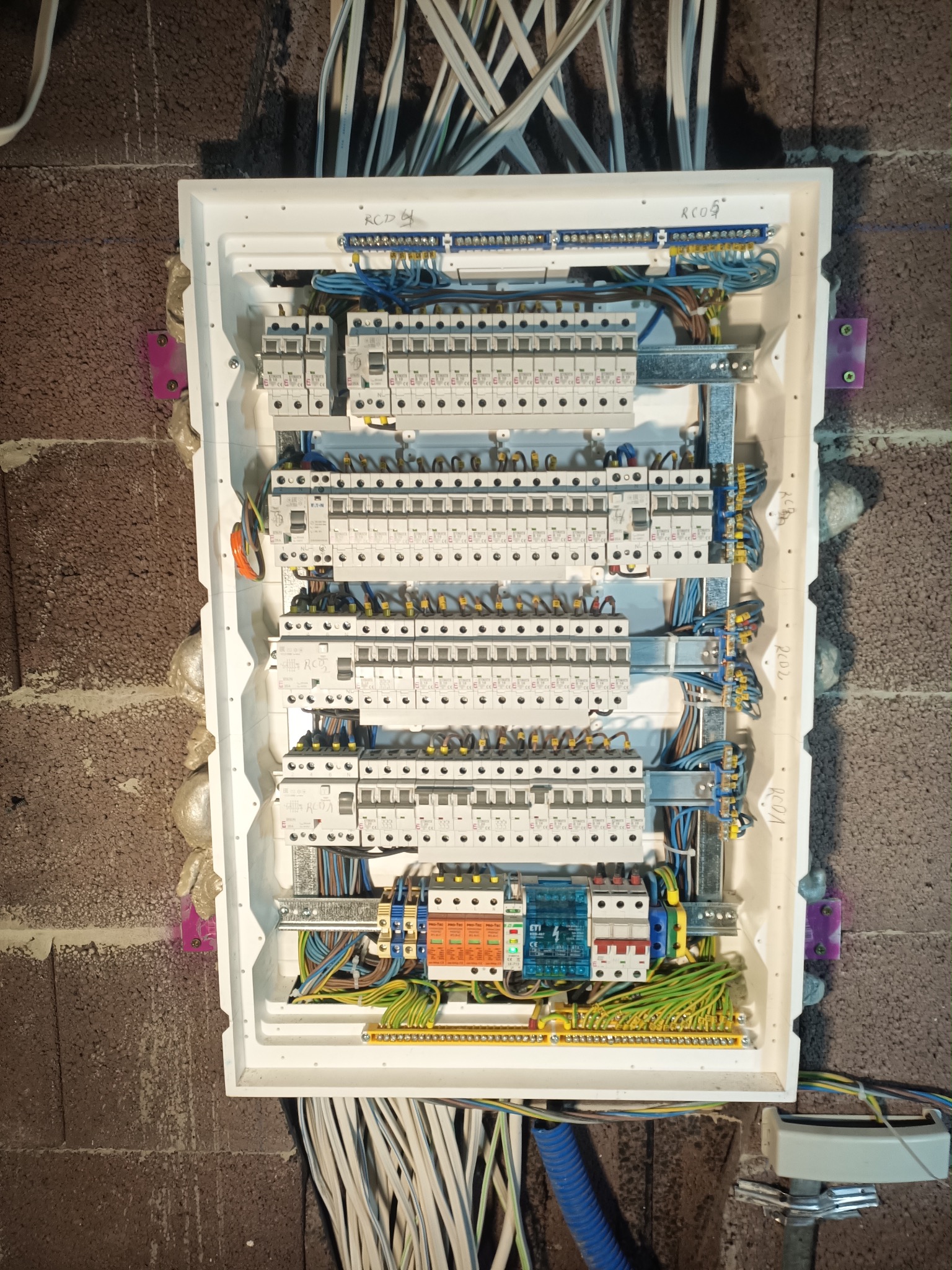Rozdzielnica elektryczna z licznymi zabezpieczeniami i okablowaniem, zamontowana na ceglanej ścianie. Widoczne bezpieczniki, przewody w różnych kolorach i oznaczenia.