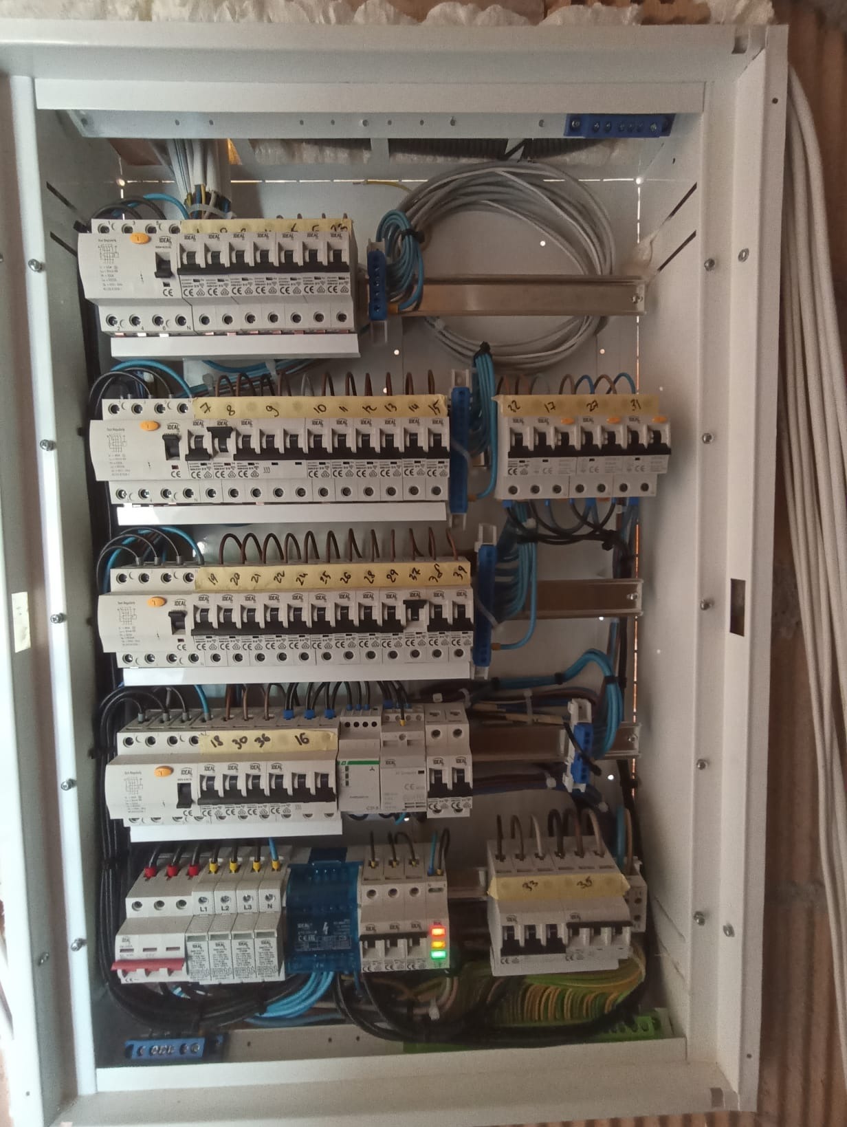 Rozdzielnia elektryczna w białej obudowie, widoczne liczne bezpieczniki, przewody i oznaczenia. Instalacja w trakcie montażu, z elementami oznaczonymi numerami.