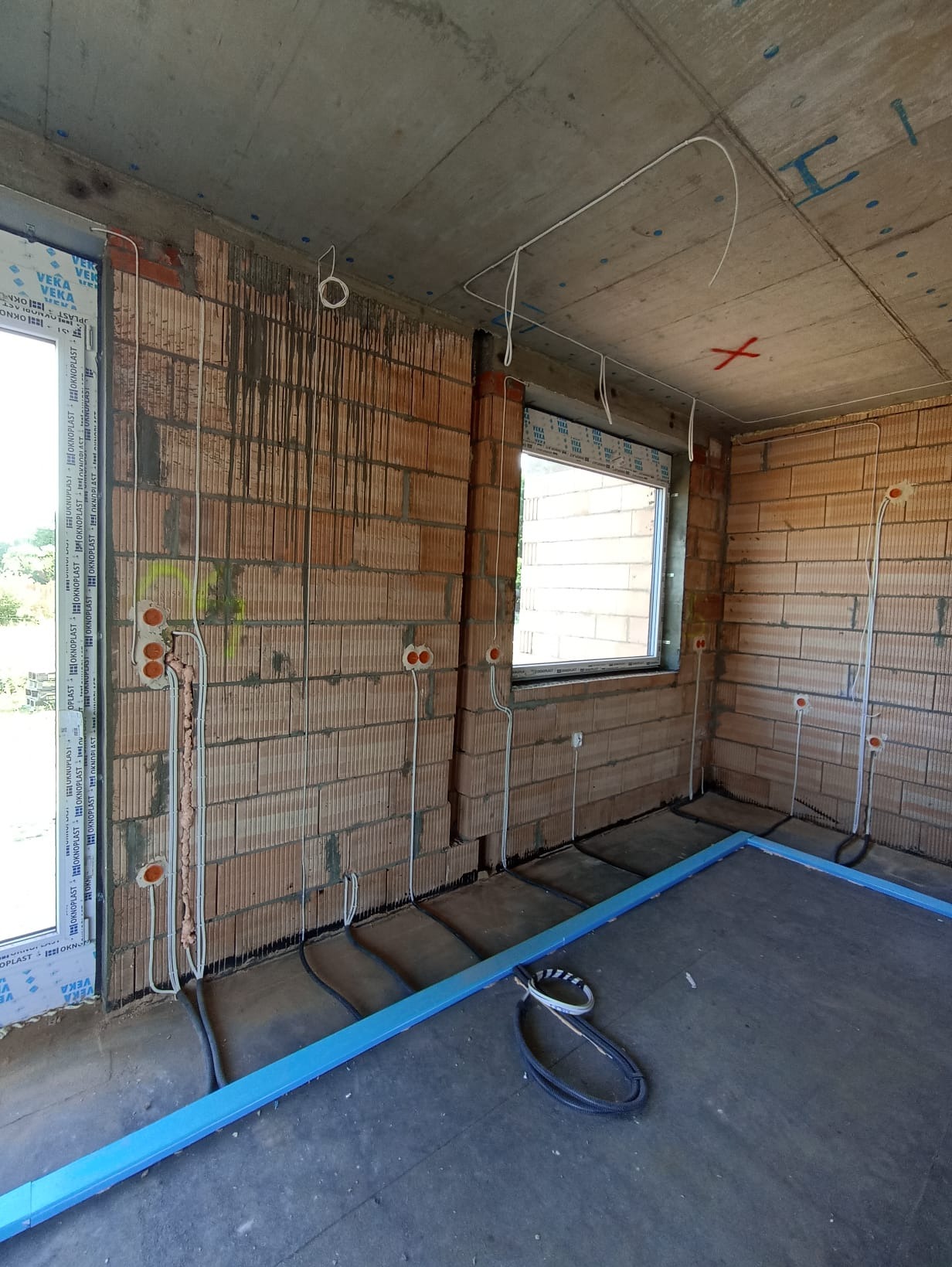 Surowy stan wnętrza z instalacją elektryczną: widoczne przewody, puszki w ścianach z cegły, okna z PCV, przygotowane podłoże pod wylewkę.