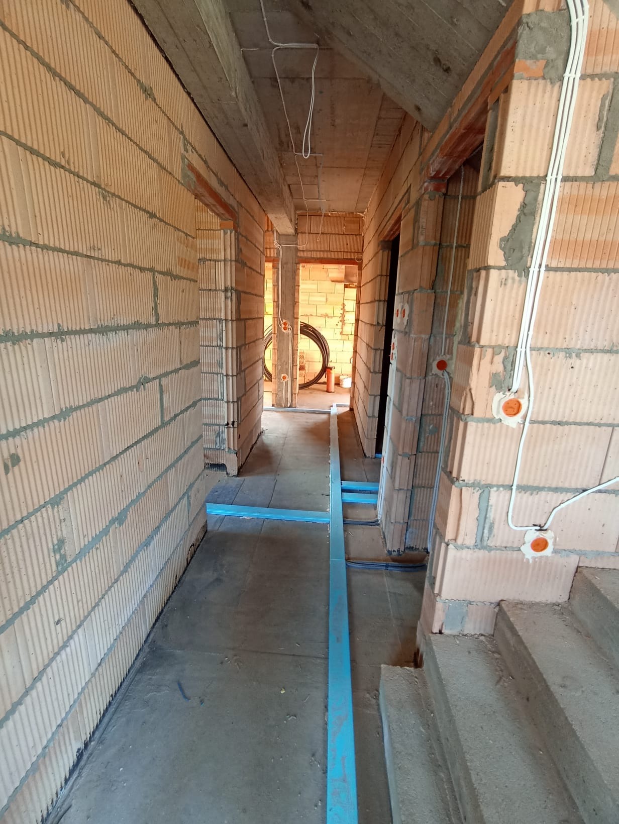 Wnętrze w trakcie budowy: ściany z cegły, instalacja elektryczna w peszlach, niebieskie profile podłogowe. Widok korytarza w surowym stanie.