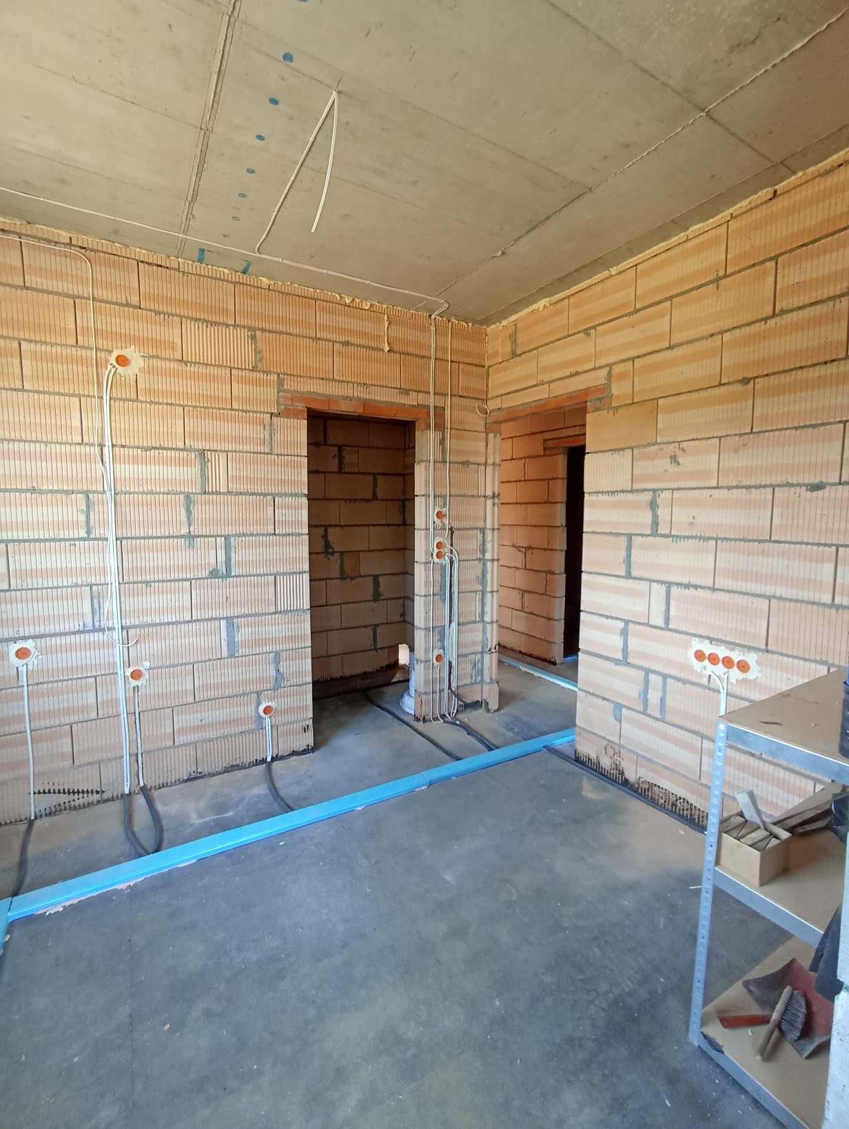 Wnętrze w trakcie prac elektrycznych: widoczne przewody w ścianach z cegły dziurawki, niebieskie rury na podłodze i puszki instalacyjne. Surowy stan wykończenia.
