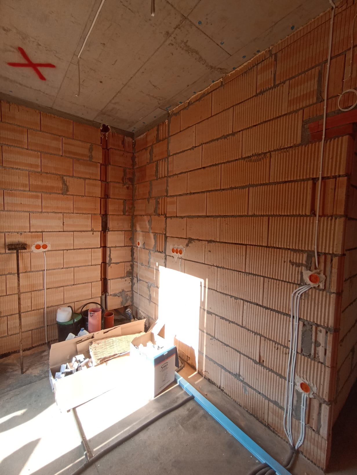 Surowy pokój z ceglanymi ścianami w trakcie instalacji elektrycznej: widoczne kable w peszlach, puszki pod gniazdka i oznaczenia na suficie. Częściowo widoczne materiały budowlane.
