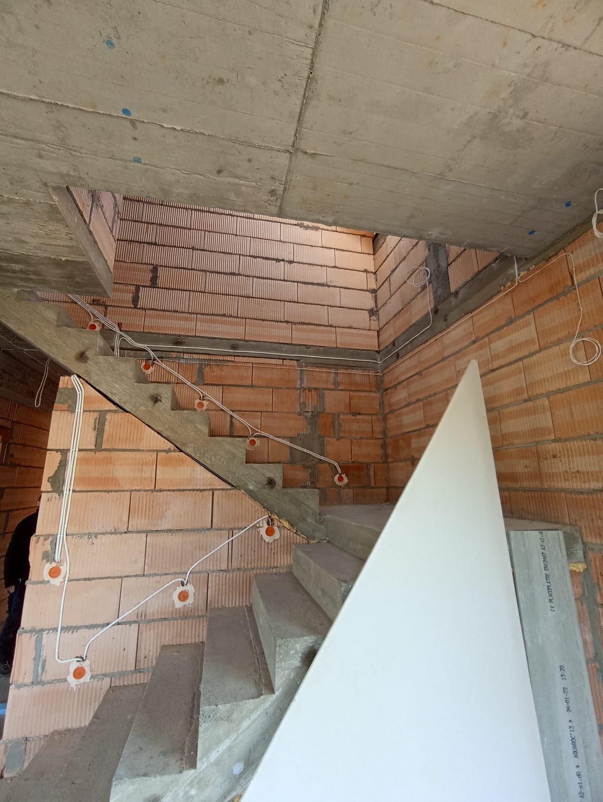 Surowe schody z ceglanymi ścianami w trakcie instalacji elektrycznej. Widoczne kable i puszki elektryczne. Częściowo widoczna płyta gipsowa. Surowy stan wykończenia.