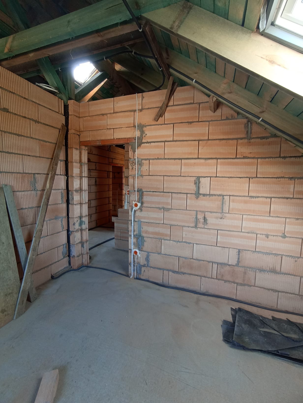 Surowy stan poddasza z oknem dachowym: ściany z cegły, widoczne przewody elektryczne w peszlach, betonowa wylewka. Etap prac wykończeniowych w nowym domu.