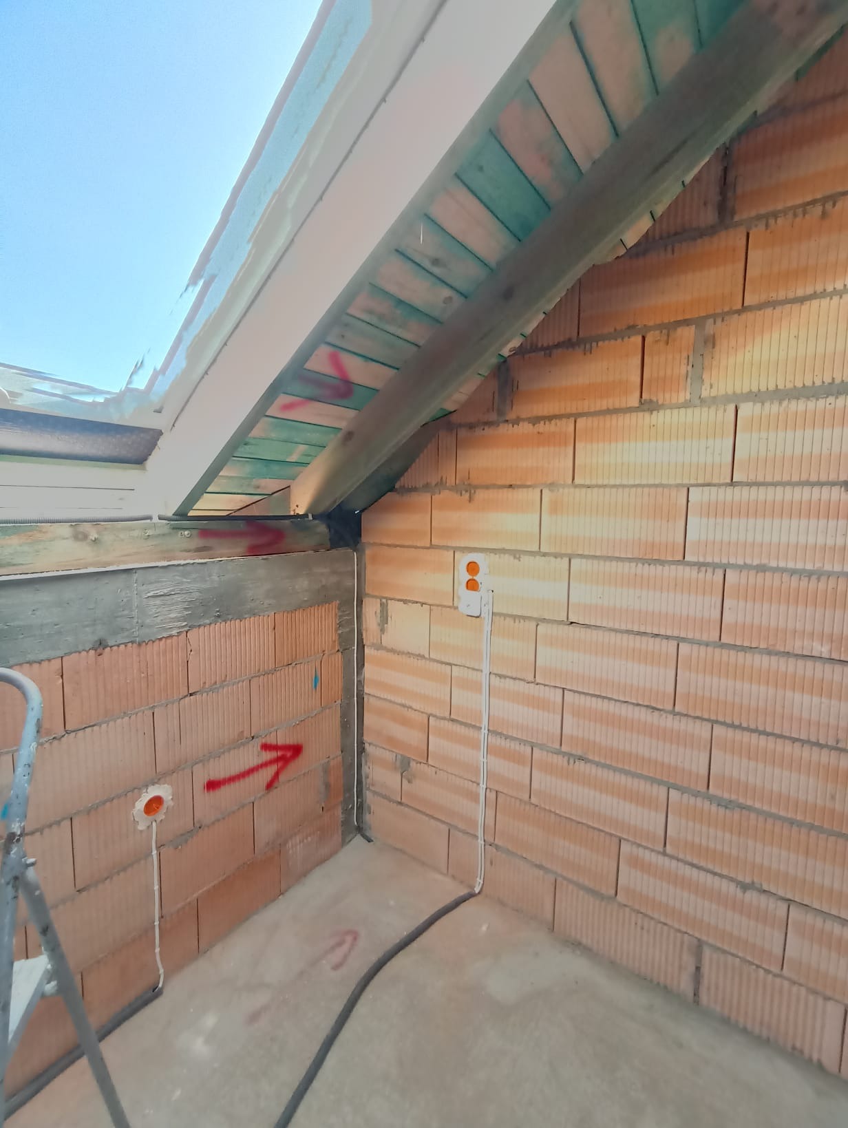 Surowy mur z instalacją elektryczną wewnątrz budynku, widoczne kable i puszki. Czerwone strzałki na ścianie. W tle drewniana konstrukcja dachu.