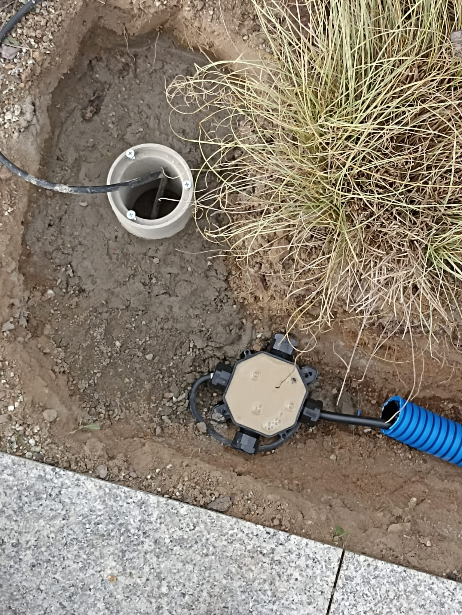 Podziemna instalacja elektryczna w wykopie: studzienka rewizyjna, puszka połączeniowa i niebieski peszel. Widoczne kable i ziemia. Prace instalacyjne w ogrodzie.