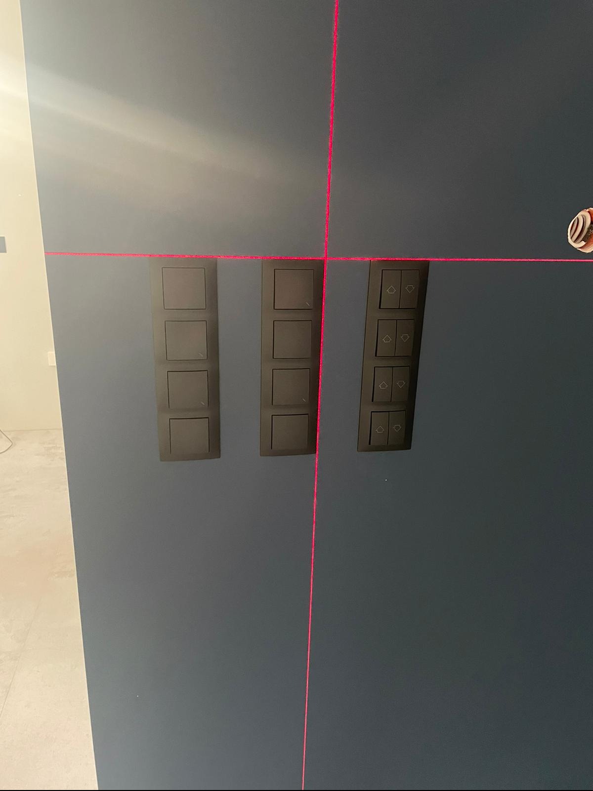 Szary panel ścienny z trzema rzędami czarnych włączników światła, wyrównanych za pomocą lasera krzyżowego. Widoczny fragment instalacji elektrycznej.