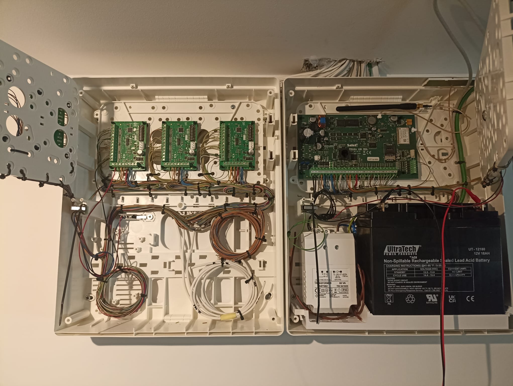 Otwarta skrzynka z elektroniką alarmową i akumulatorem w Szczecinie. Widoczne płytki drukowane, przewody i transformator. Instalacja zabezpieczeń wewnątrz obudowy.