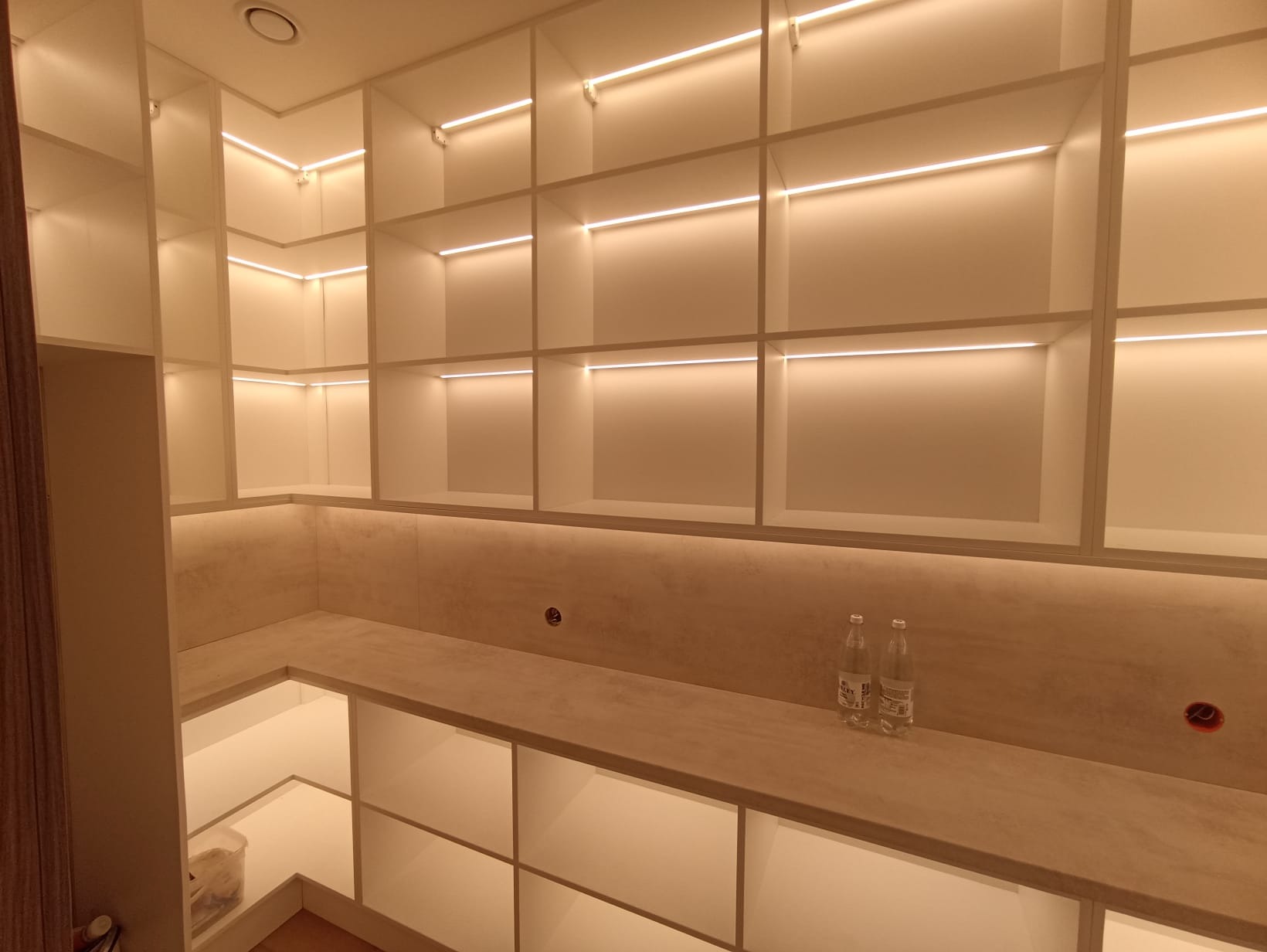 Biała, podświetlana półka wnękowa z blatem, dwie butelki wody na blacie. Oświetlenie LED podkreśla nowoczesny design. Widoczne gniazdka elektryczne.