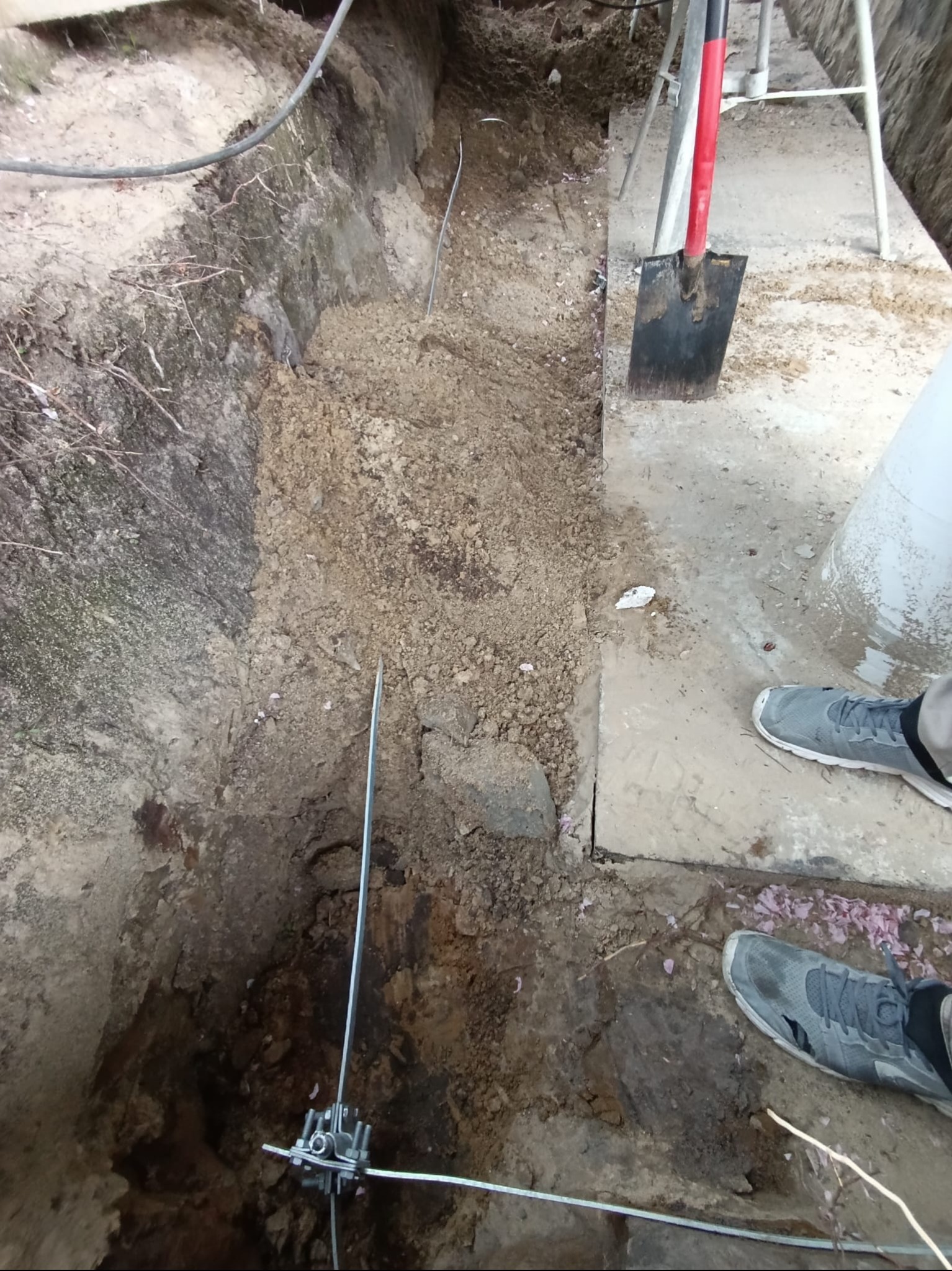 Wykop z ziemią, w którym widoczne są metalowe pręty i złącza, prawdopodobnie element instalacji odgromowej. Obok leży szpadel i widać fragment buta.