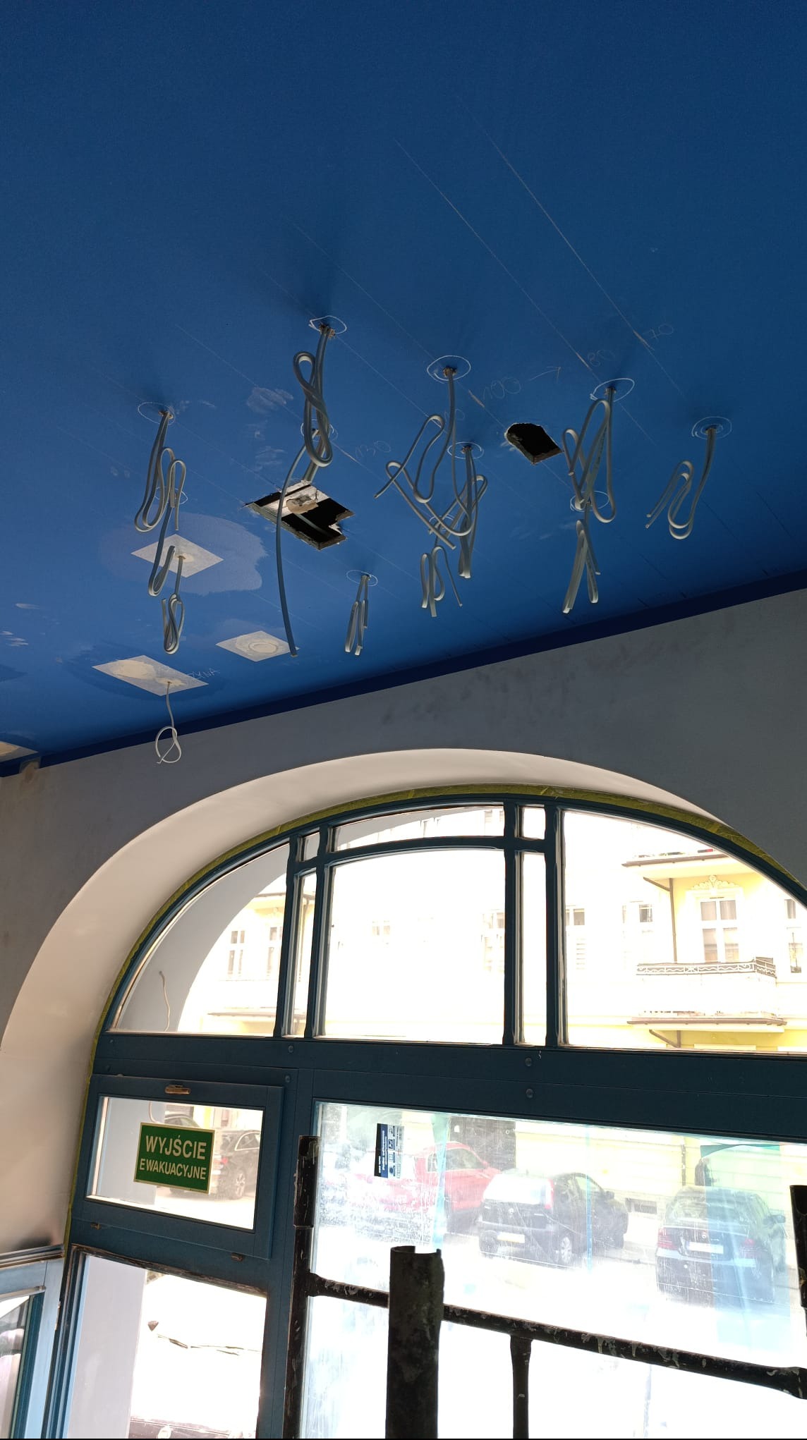 Niebieski sufit z wystającymi przewodami elektrycznymi w trakcie instalacji, widoczne otwory montażowe i okno z napisem 'Wyjście Ewakuacyjne'.
