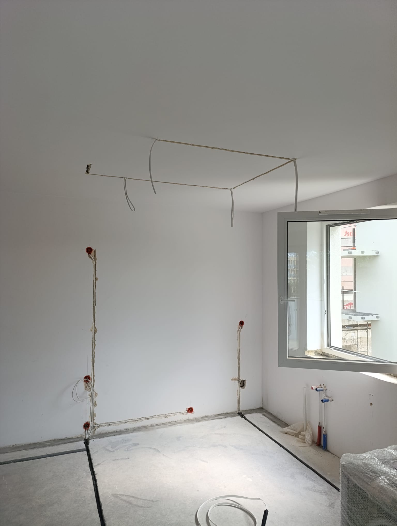 Wnętrze w trakcie prac elektrycznych: widoczne kable w ścianach i na suficie, puszki elektryczne, surowa podłoga i okno z widokiem na zewnątrz. Etap instalacji elektrycznej.