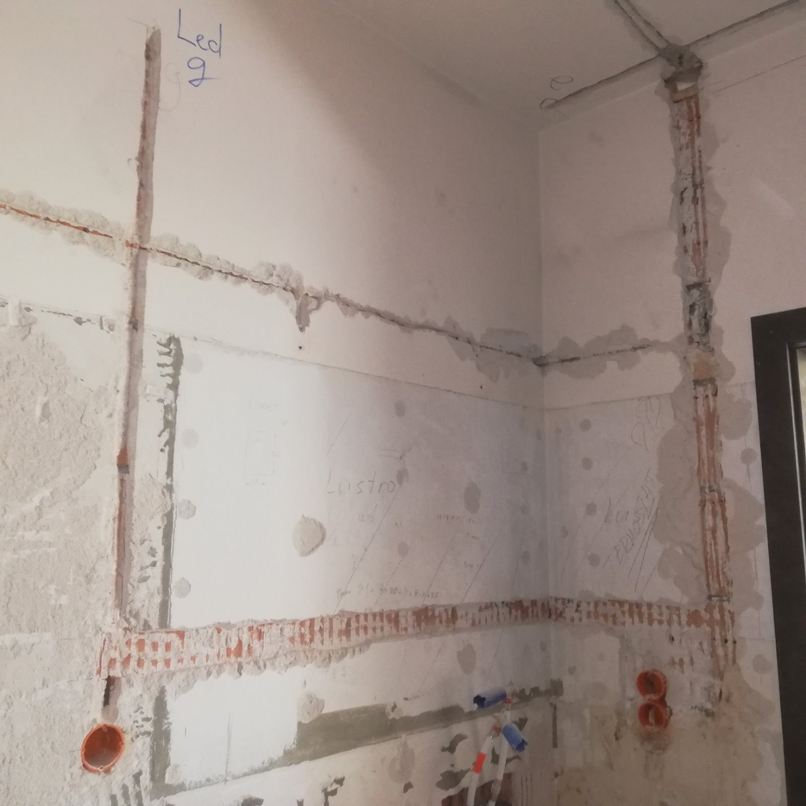Ściana w trakcie remontu z widocznymi przewodami elektrycznymi w bruzdach, puszkami instalacyjnymi i notatkami ołówkiem na ścianie.