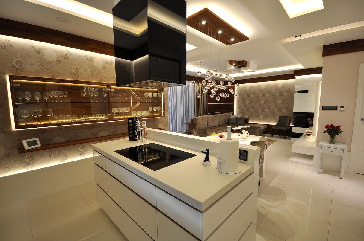 Nowoczesna kuchnia z wyspą i przeszkloną szafką z podświetleniem LED. W tle salon z sofą i dekoracyjnym oświetleniem. Całość w jasnej kolorystyce z drewnianymi akcentami.