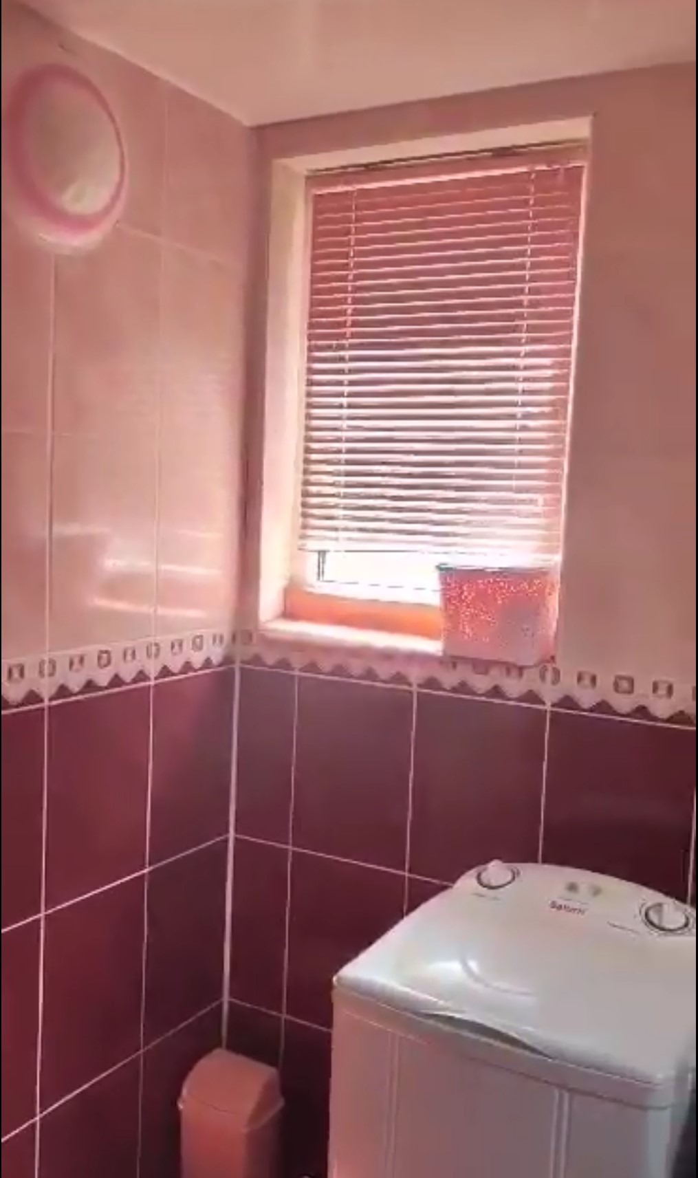 Wykończona łazienka w odcieniach różu i bordo z pralką, okrągłym lustrem i żaluzjami w oknie. Widoczne płytki na ścianach i dekoracyjny pasek.