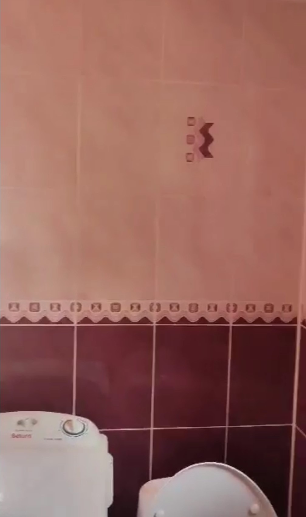 Wykończona łazienka z bordowymi i beżowymi płytkami, dekoracyjnym pasem i wzorem na ścianie. Widoczny fragment toalety i pralki.