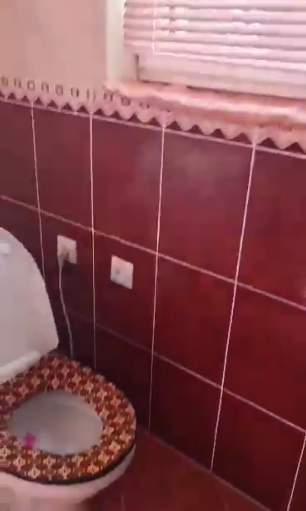 Czerwona, kwadratowa glazura na ścianach i podłodze łazienki. Widoczny fragment toalety z wzorzystą deską i okno z żaluzjami. Gniazdka elektryczne na ścianie.