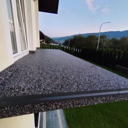 Green House - Wykończony taras z szarego kamienia kompozytowego, widok z boku, z oknem i fragmentem elewacji domu. W tle zieleń drzew i górski krajobraz o zmierzchu.