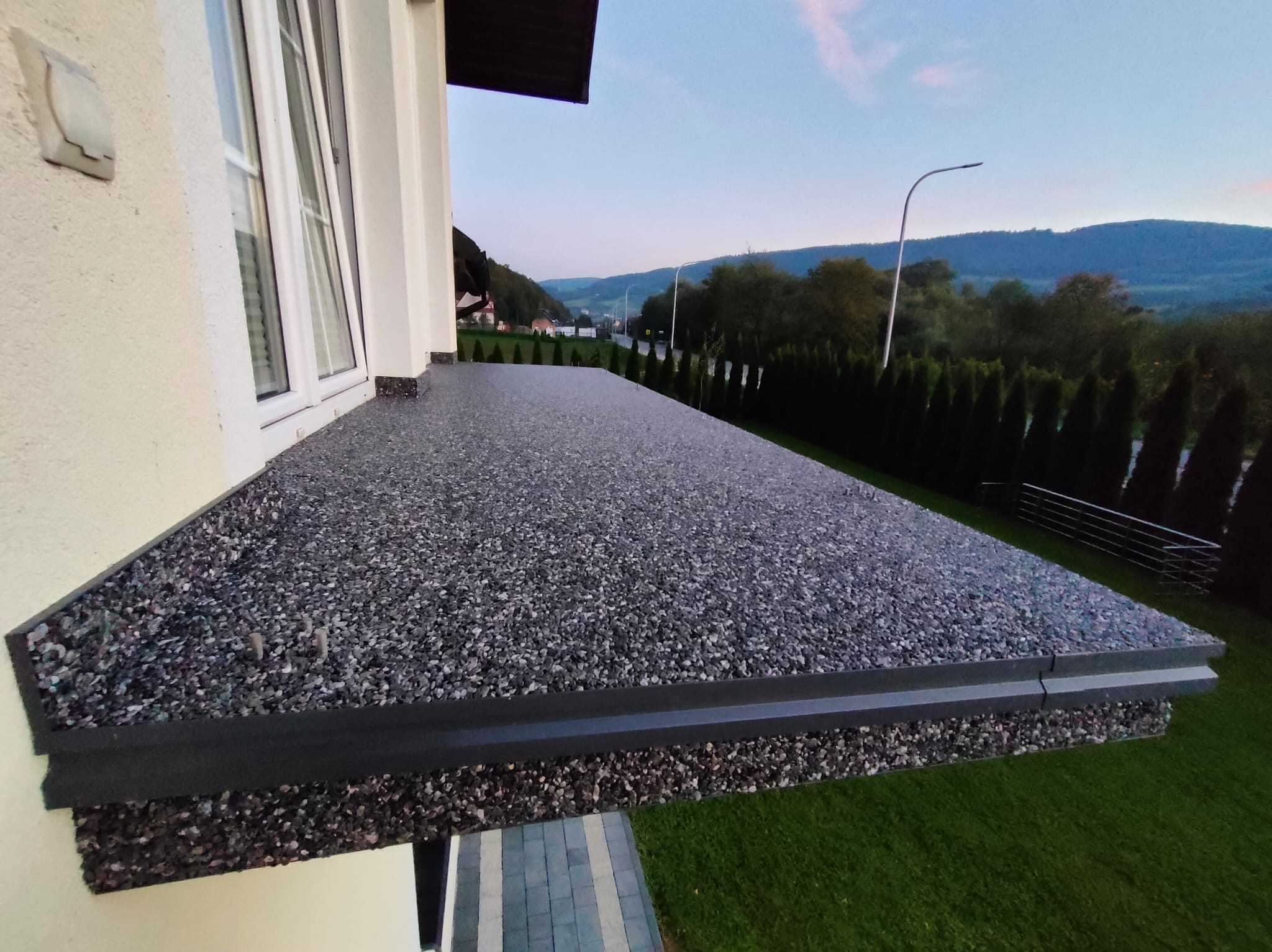 Wykończony taras z szarego kamienia kompozytowego, widok z boku, z oknem i fragmentem elewacji domu. W tle zieleń drzew i górski krajobraz o zmierzchu.