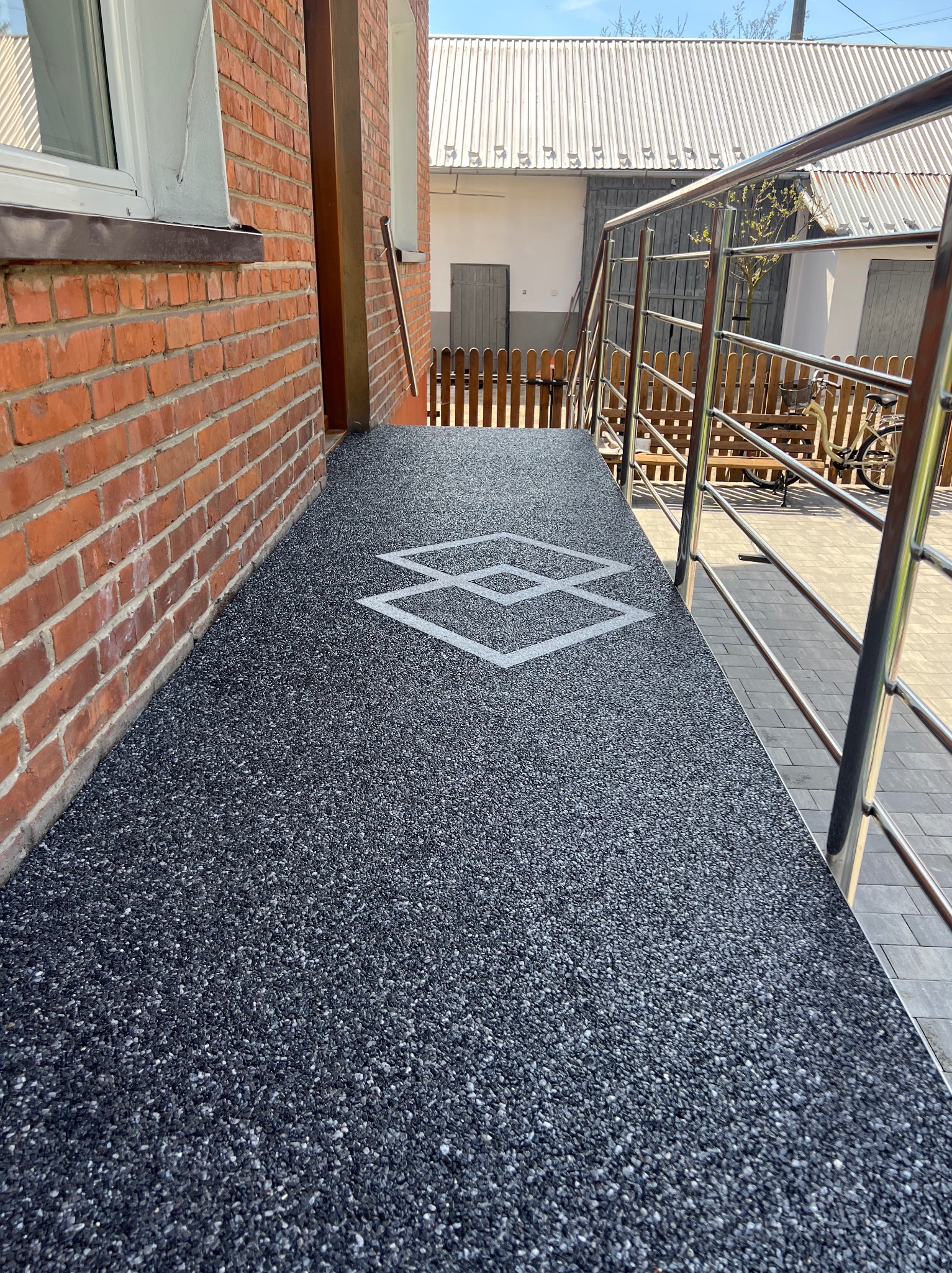 Wykończony taras kamienny z żywicy epoksydowej w kolorze grafitowym, z geometrycznym wzorem. Widoczne metalowe barierki i fragment elewacji z cegły.