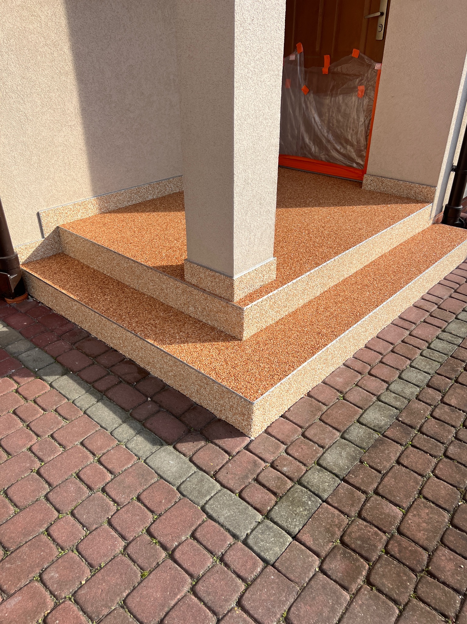 Wykończone żywicą schody wejściowe z beżową kolumną i kostką brukową. Zabezpieczone drzwi wejściowe folią malarską. Widok z góry na detale wykończenia.