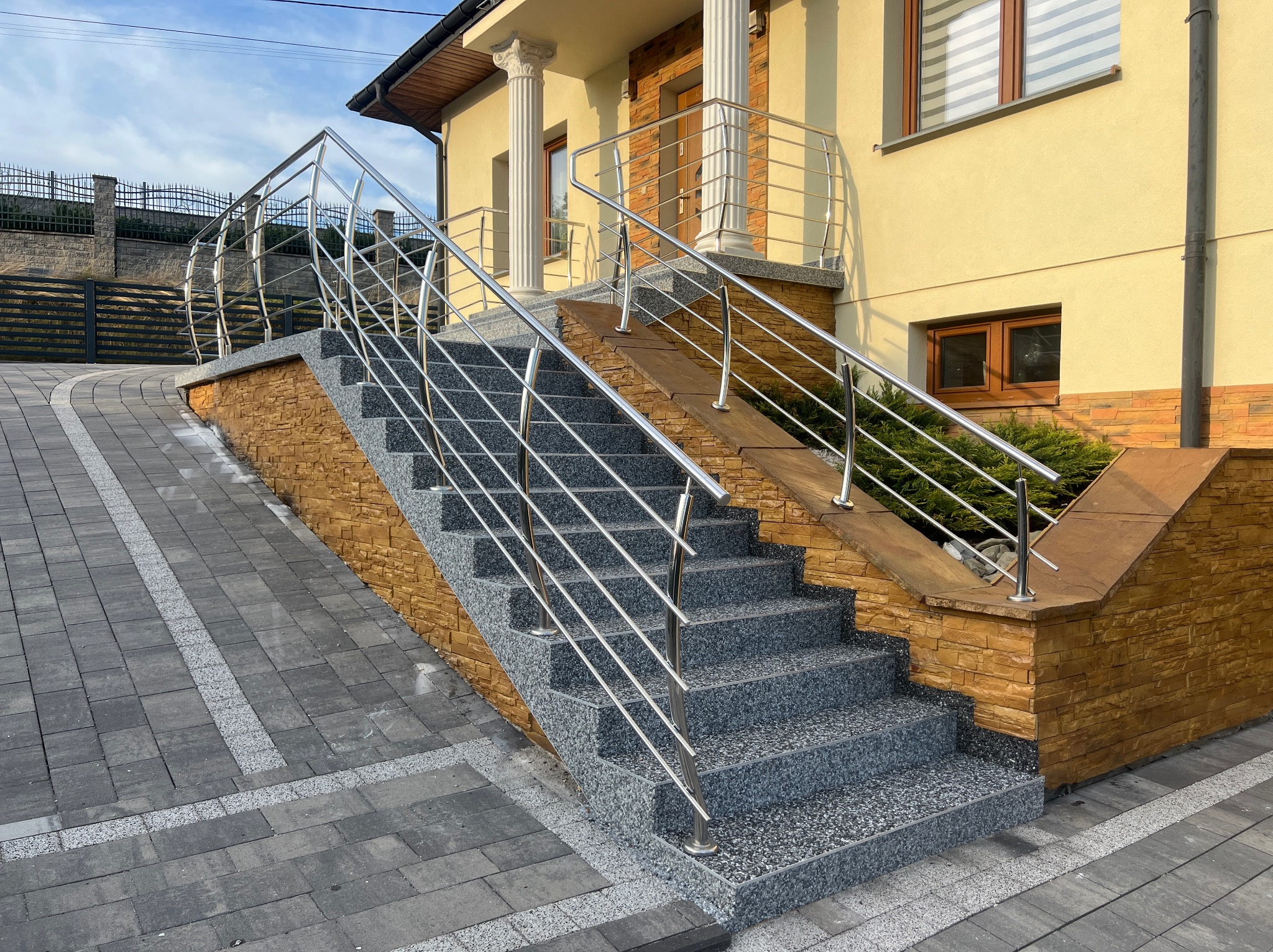 Zewnętrzne schody z szarego kamienia z metalową balustradą prowadzące do domu z kolumnami. Ułożona kostka brukowa na podjeździe.