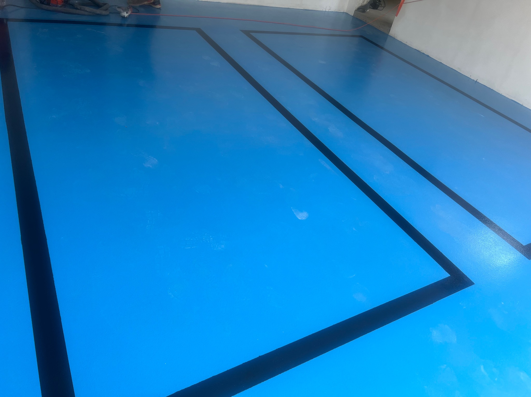 Błękitna podłoga z czarnymi liniami tworzącymi geometryczny wzór. Widoczne ślady stóp i fragment sprzętu budowlanego w tle. Nowoczesne wykończenie podłogi.