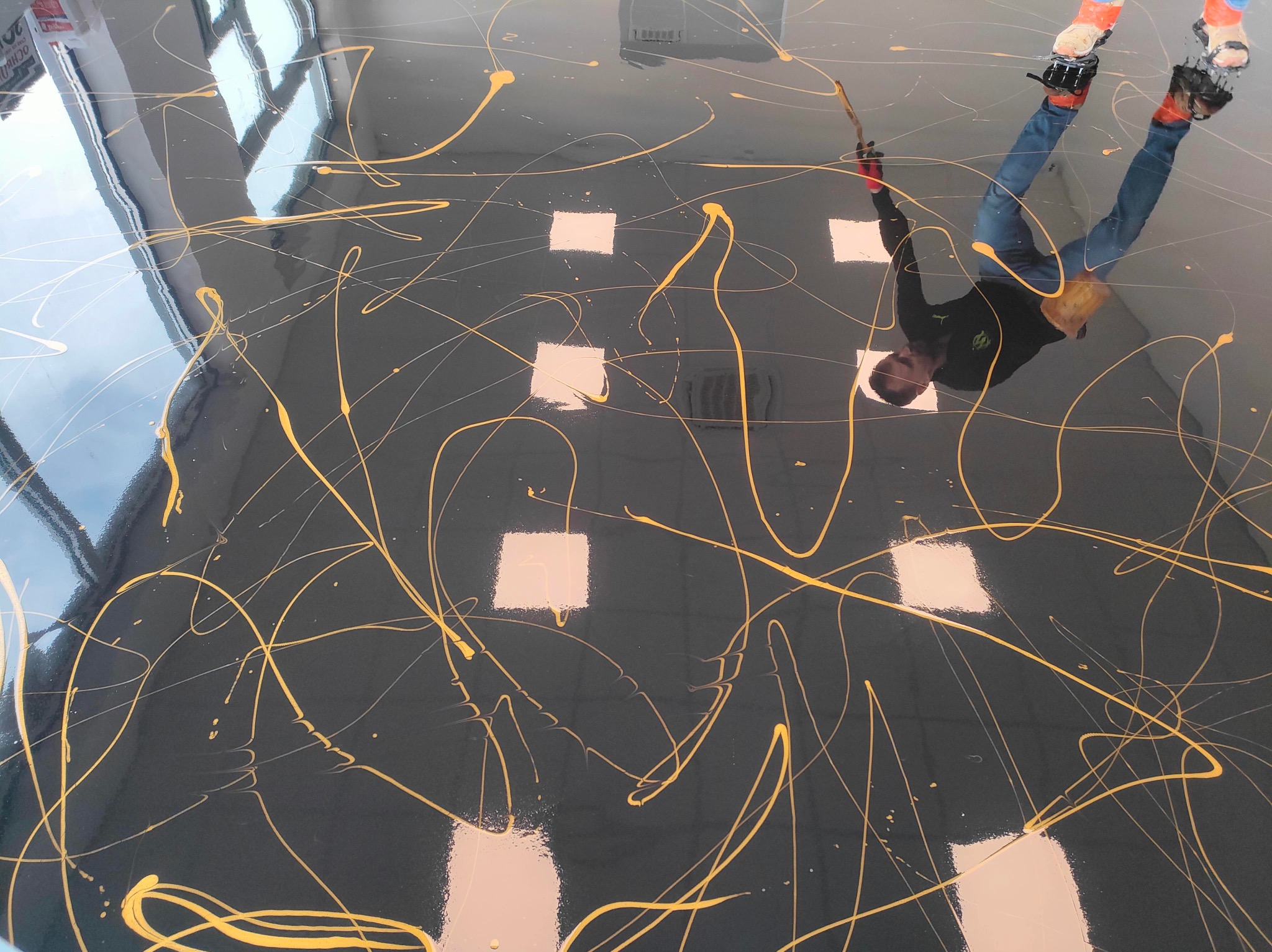 Błyszcząca, ciemna posadzka z abstrakcyjnym, złotym wzorem. Widoczne odbicie osoby pracującej nad wykończeniem podłogi. Nietypowe ujęcie z góry.