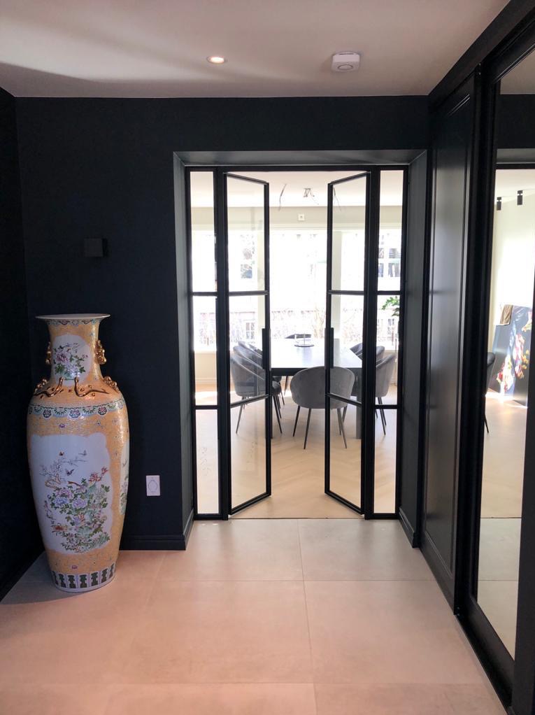 Czarne, przeszklone drzwi wewnętrzne w stylu loft, otwarte do jadalni z szarymi krzesłami. Duży, zdobiony wazon stoi obok wejścia na jasnej podłodze.