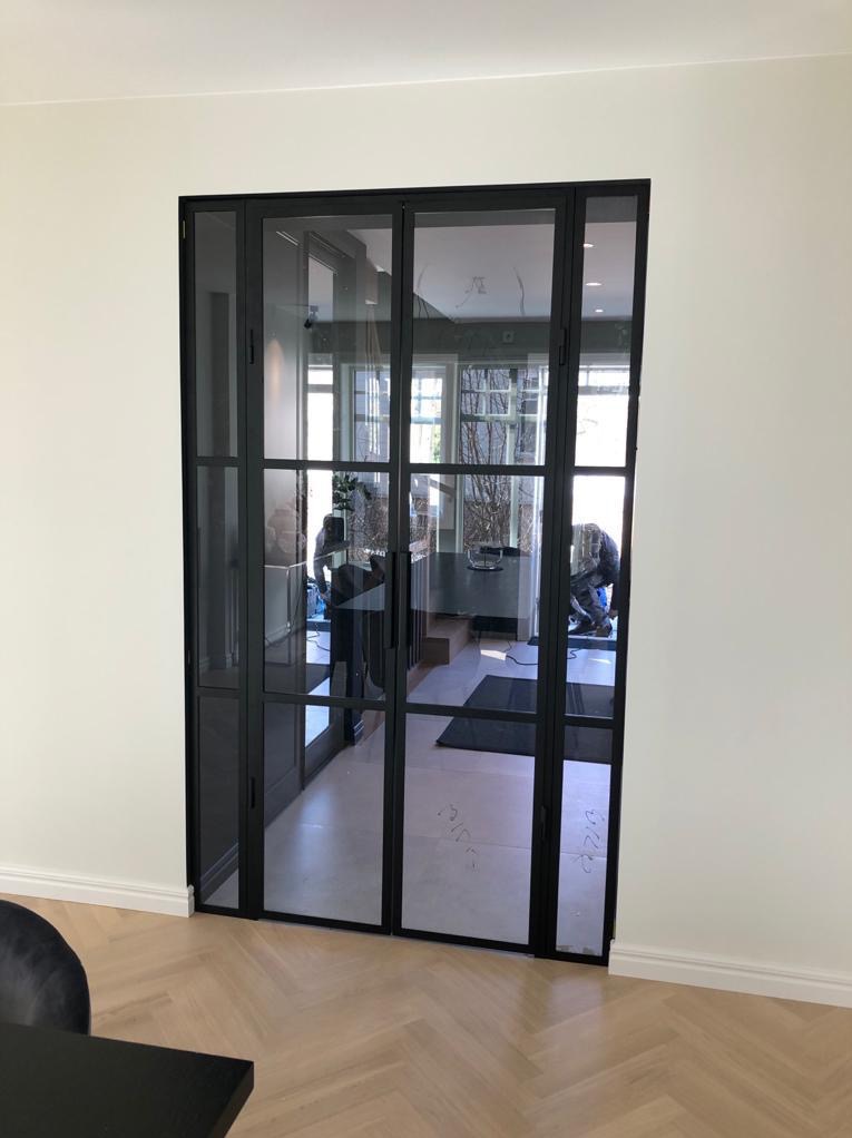Nowoczesne, czarne drzwi wewnętrzne z przeszkleniami, dzielone szprosami, w jasnym wnętrzu z parkietem ułożonym w jodełkę. Widoczne odbicia w szybach.
