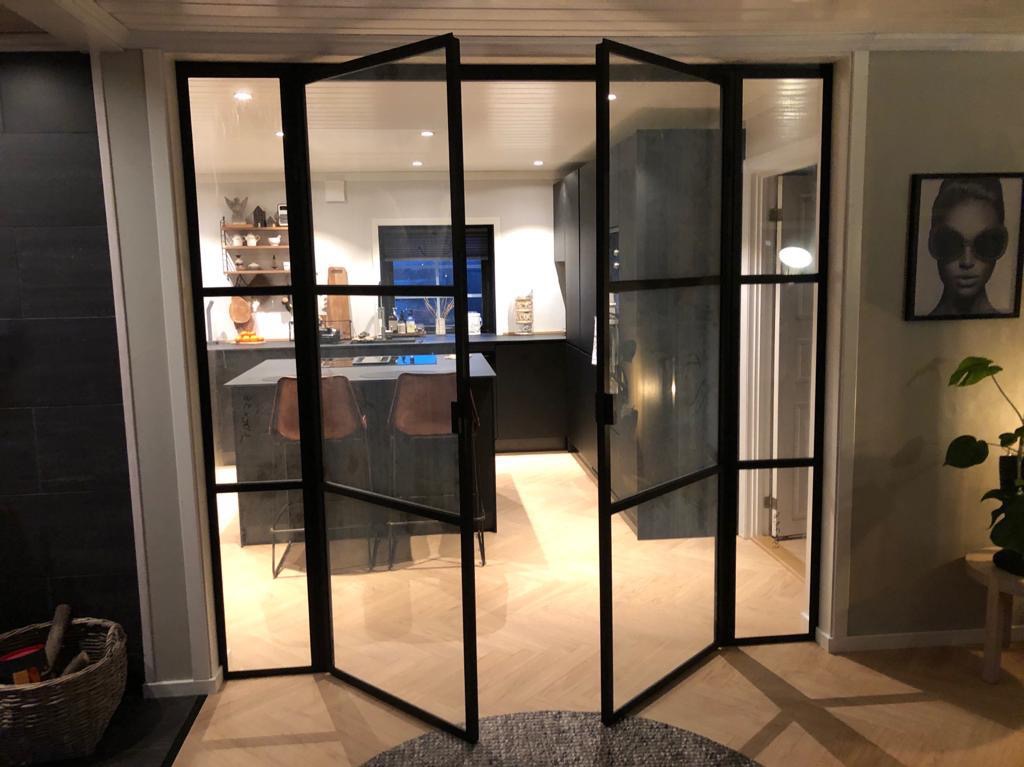 Nowoczesne, dwuskrzydłowe drzwi szklane z czarnymi szprosami, otwarte do wnętrza. Widoczna kuchnia z wyspą i krzesłami barowymi. Parkiet w jodełkę na podłodze.