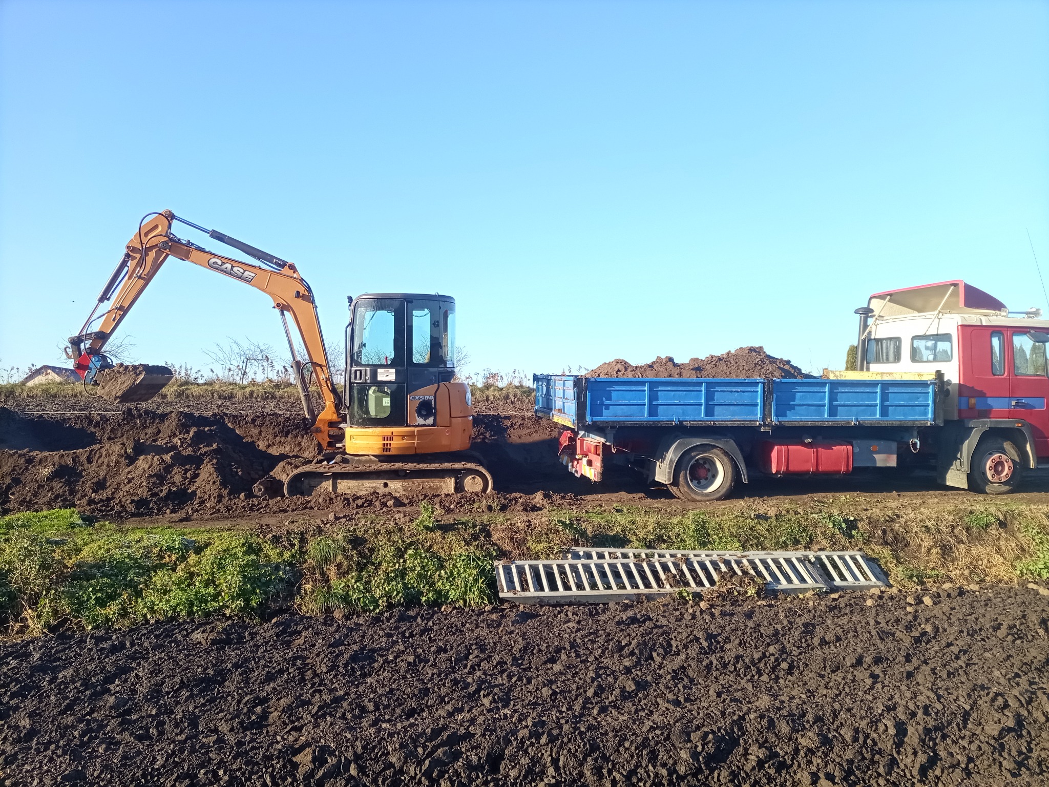Pomarańczowa koparka CASE nabiera ziemię i ładuje na niebieską ciężarówkę na tle błękitnego nieba. Widoczny fragment wykopu i metalowa krata.