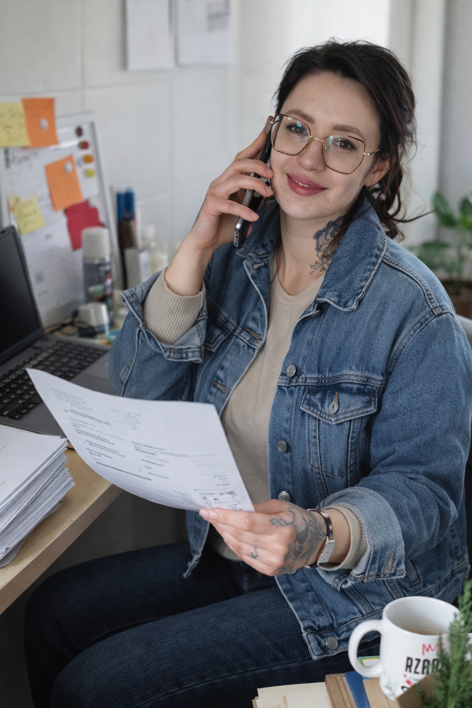 Kobieta w okularach rozmawia przez telefon, trzymając dokument. Na biurku laptop, notatki i kubek. Naturalne światło, domowe biuro.