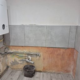 Daniel Makowski - Remont łazienki w trakcie: ułożone szare płytki podłogowe imitujące drewno i częściowo położone płytki ścienne. Widoczne rury i wiadro z zaprawą.