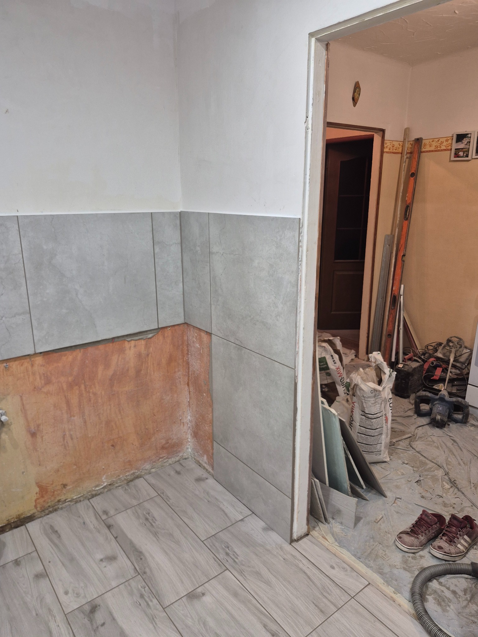 Układanie szarych płytek na ścianie i podłodze w łazience. Częściowo wykończona ściana, widoczne narzędzia i materiały budowlane. W tle otwarte drzwi do innego pomieszczenia.